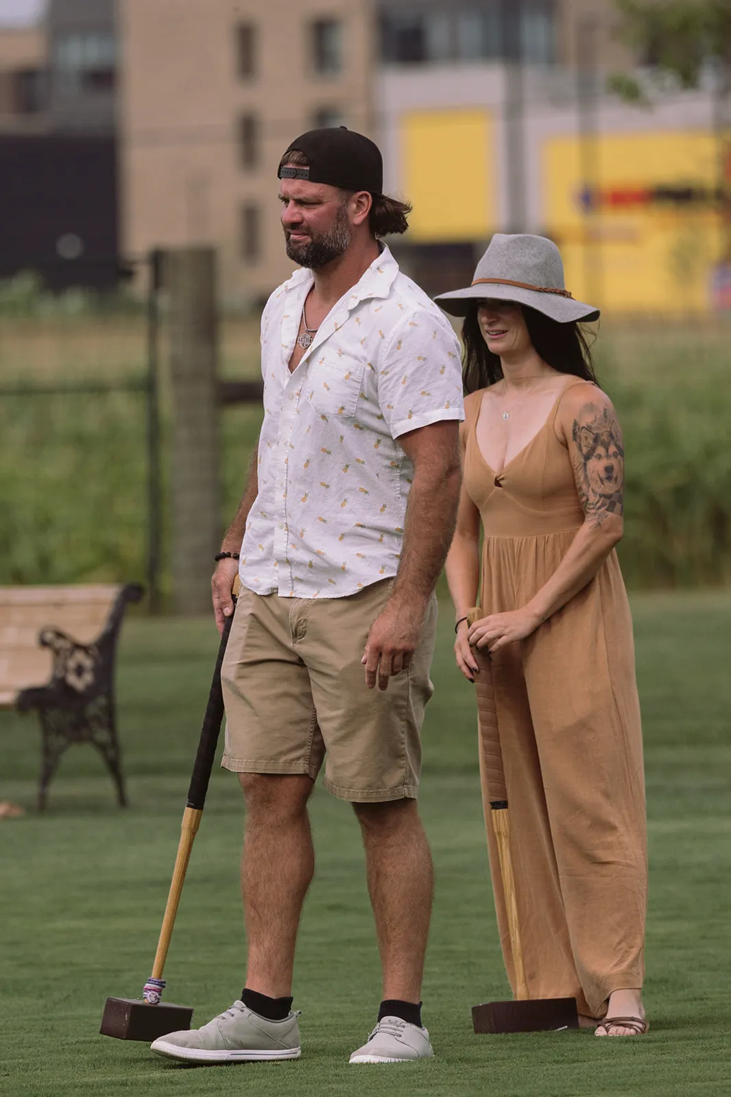 A man holding a croquet mallet and a woman in a wide-brimmed hat and tan dress stand together on the lawn during a game of croquet.
