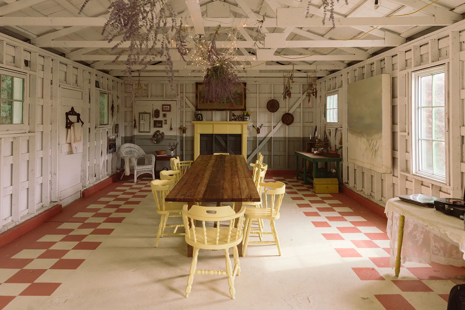 The interior of a rustic white-panelled barn set for a meal — a long wooden table ringed by pale-yellow chairs on a red-and-white checkered floor, dried lavender hanging overhead.