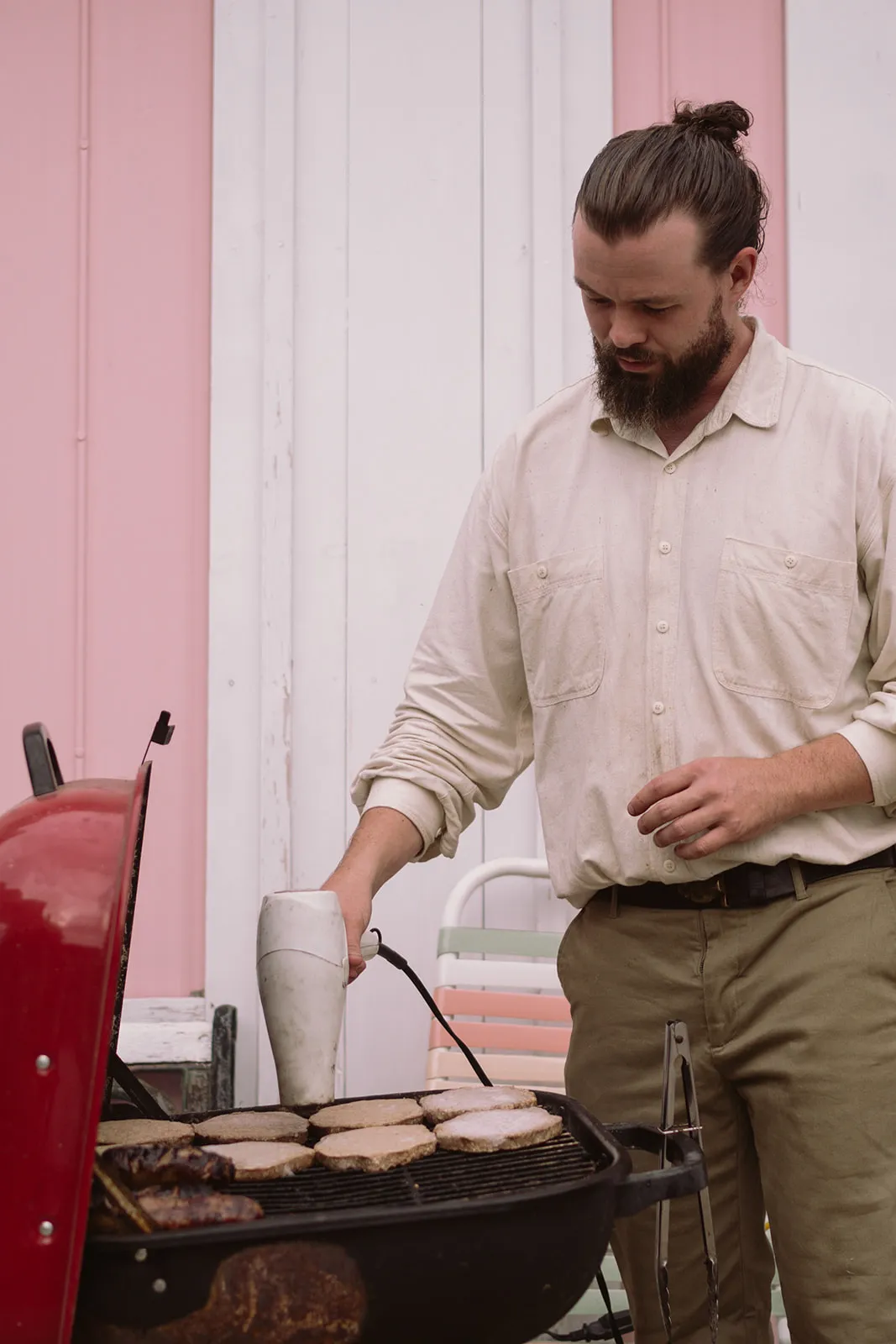 A bearded man in a cream shirt grills burgers and steaks on a red charcoal barbecue against a pink-and-white panelled wall.
