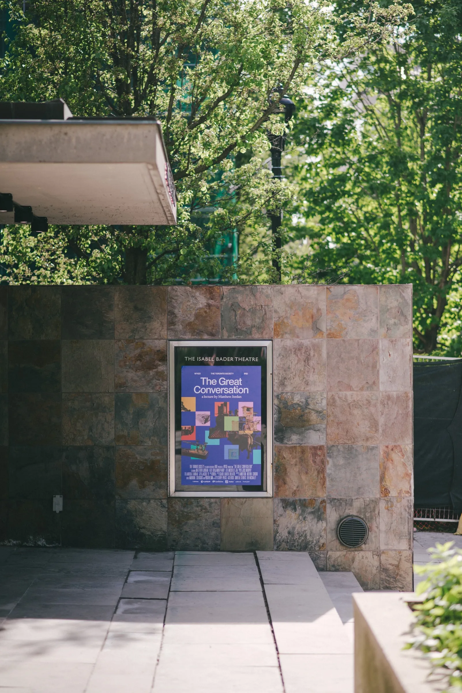 The exterior stone wall of the Isabel Bader Theatre with a framed poster for The Great Conversation, amid summer trees.