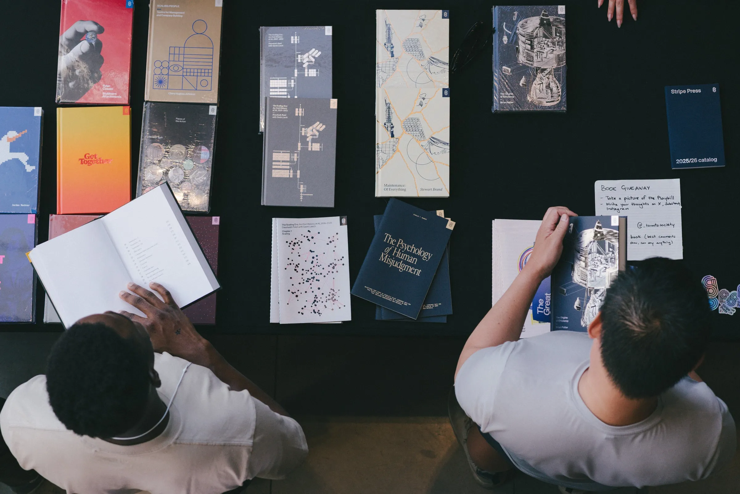 An overhead view of two attendees in white T-shirts browsing a long table of design books and a “Book Giveaway” card.