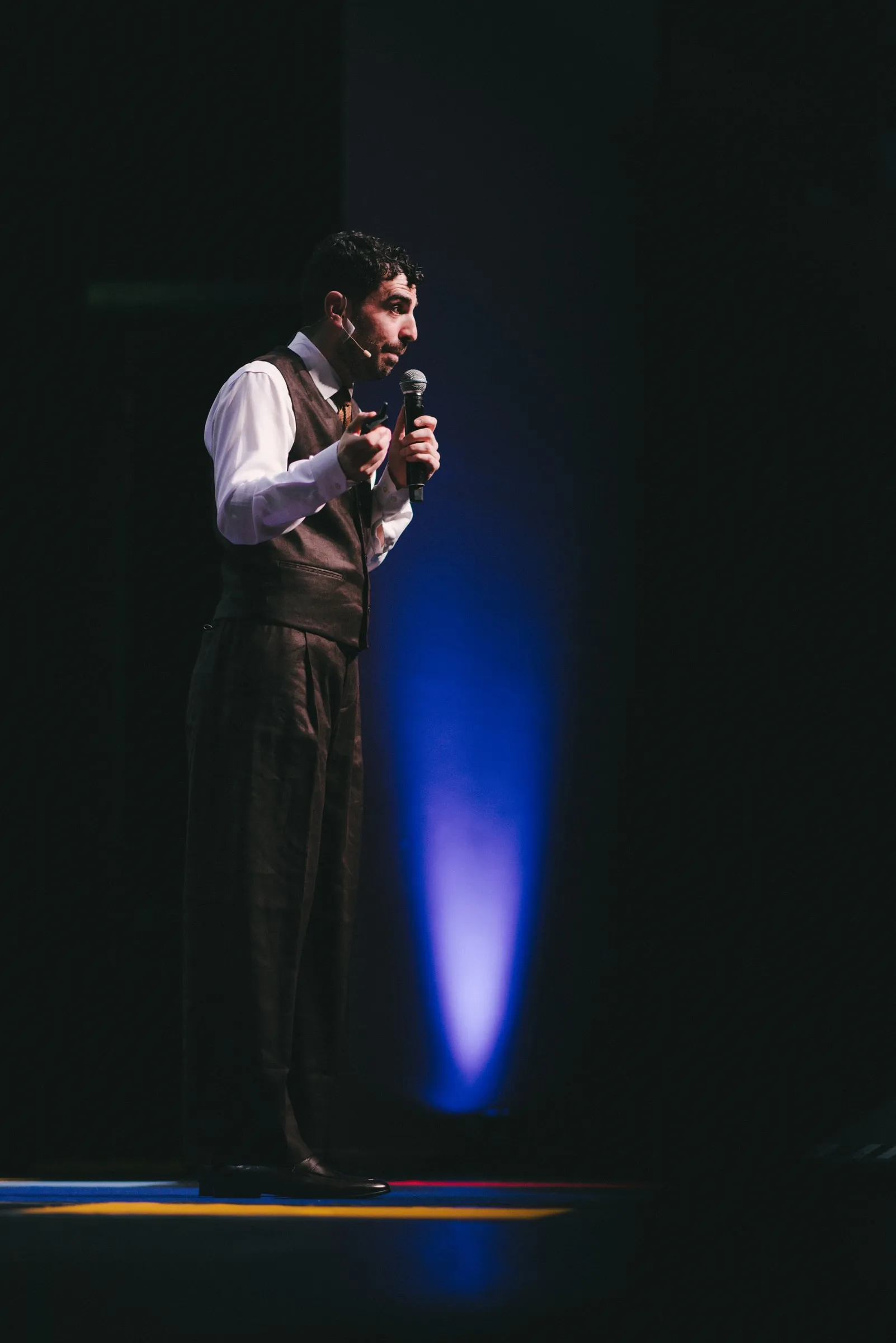 A speaker in a waistcoat and tie addresses the audience with a handheld microphone, lit by a blue stage light on the dark Isabel Bader Theatre stage.