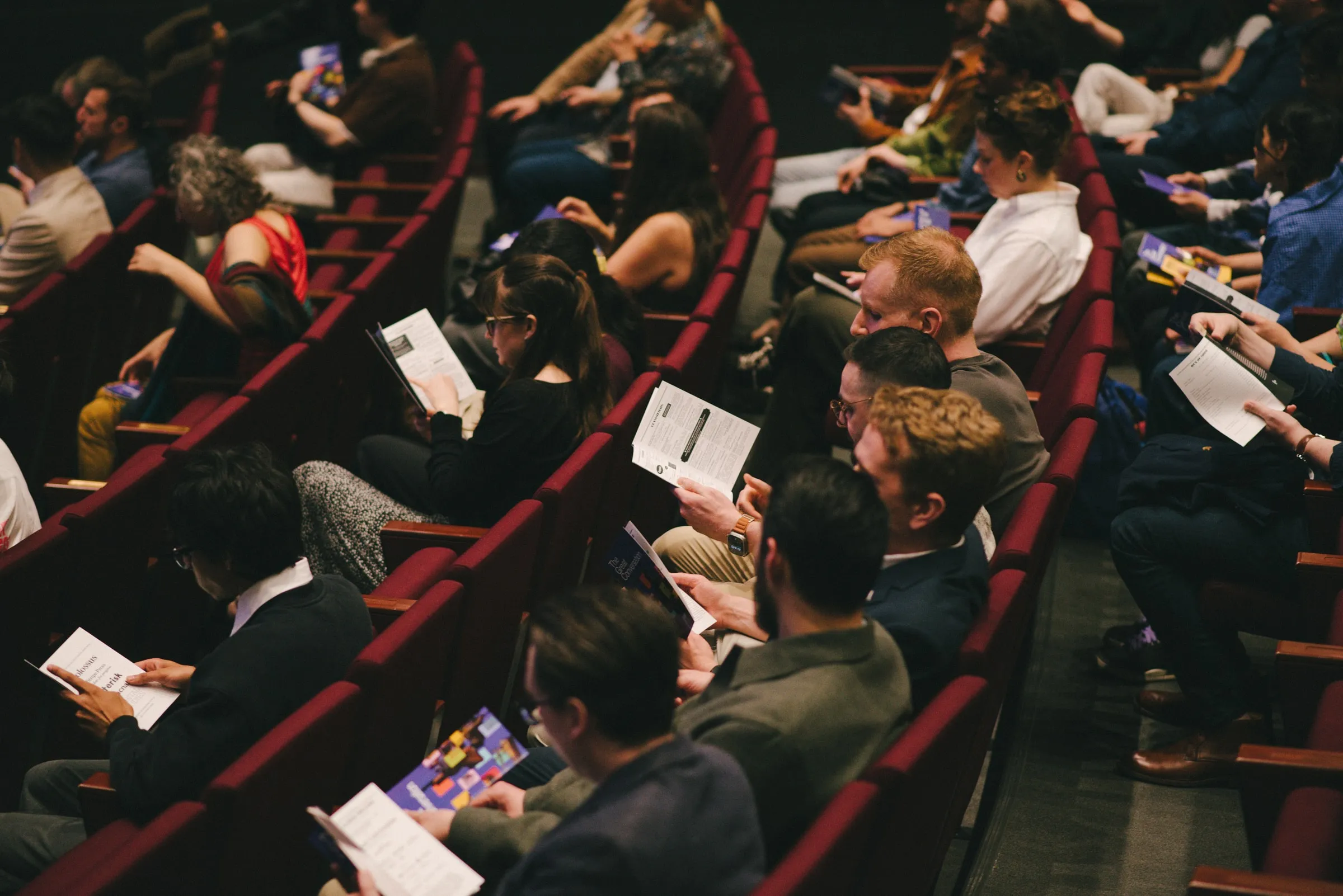 Audience members sit in the theatre’s curved rows reading the event programme.