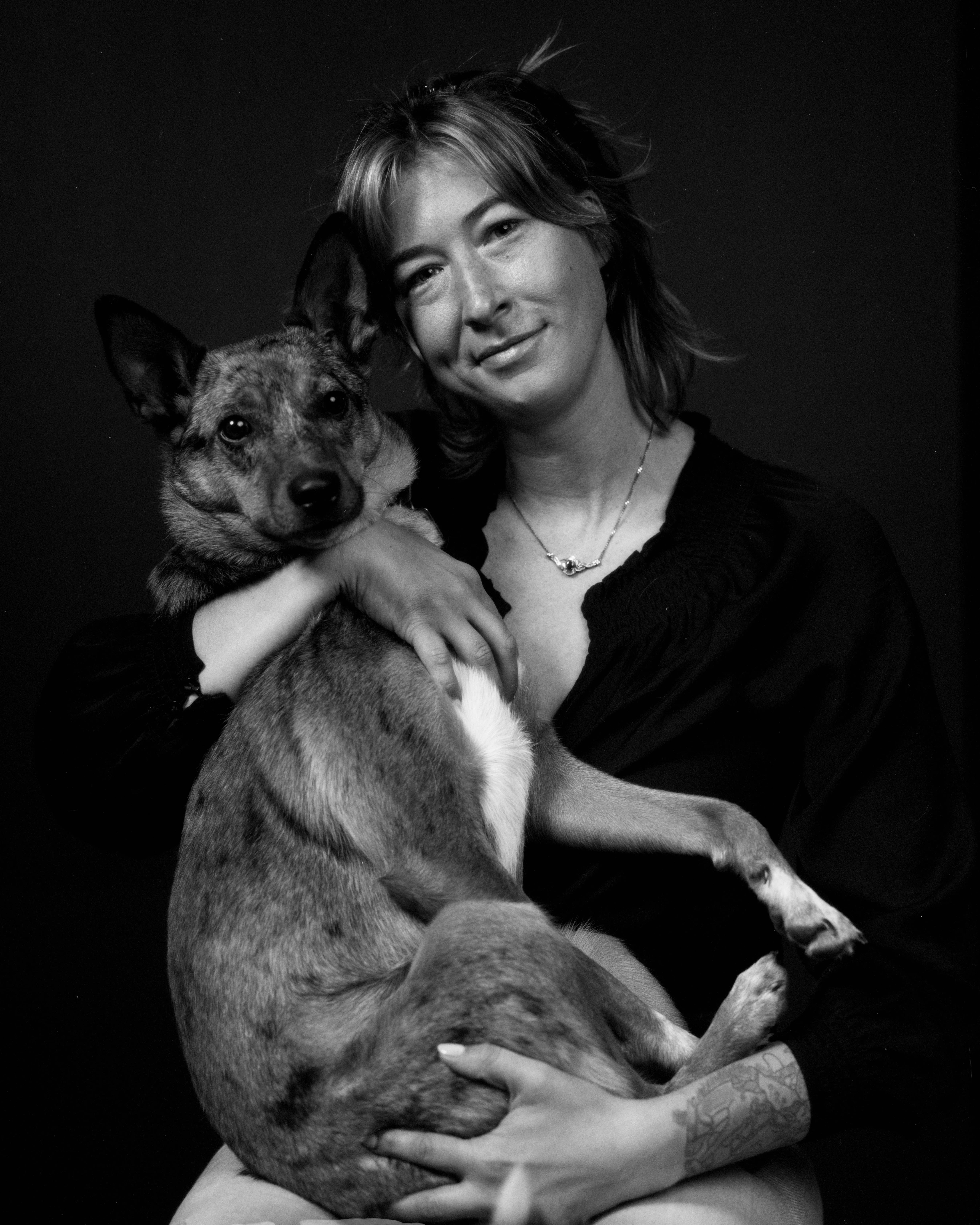 A black-and-white studio portrait of Rachel Long smiling softly, holding her cattle dog Loretta Lynn in her arms against a dark backdrop.