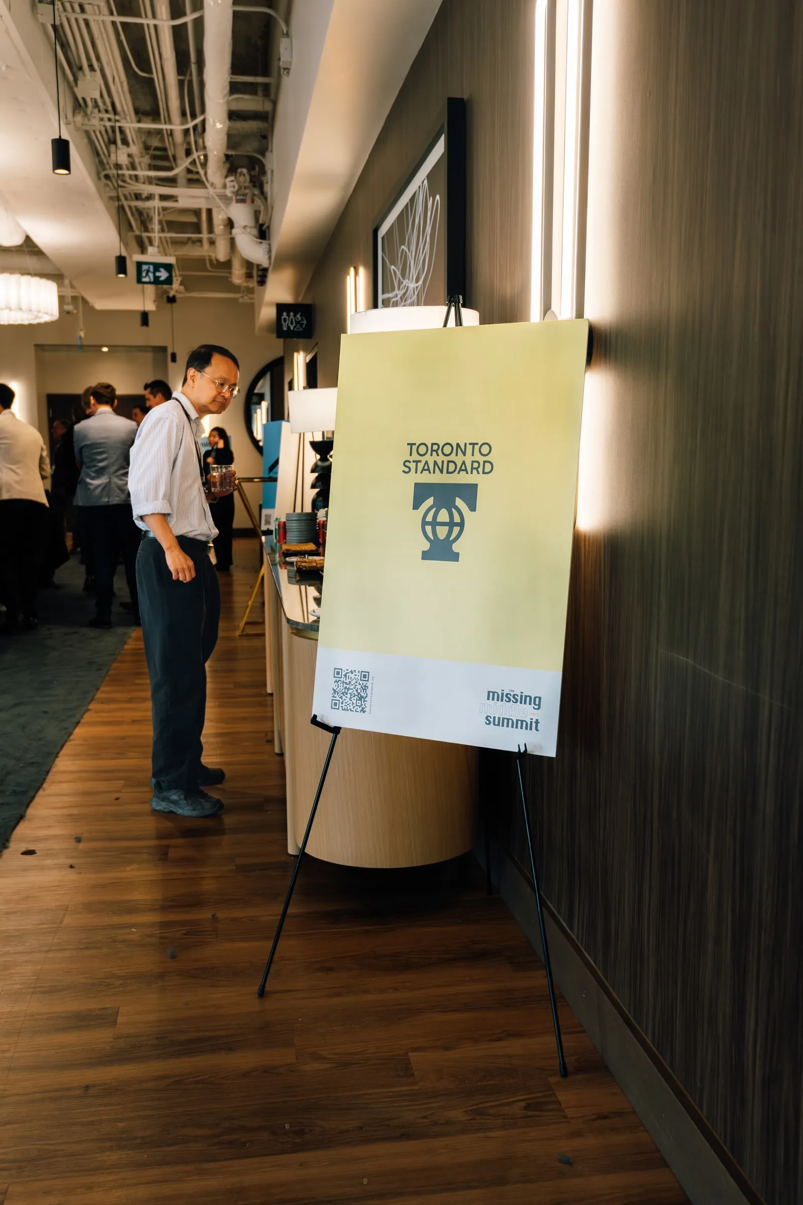 An attendee browses a refreshments counter beside a Toronto Standard easel sign for the Missing Middle Summit, in a wood-floored loft space.