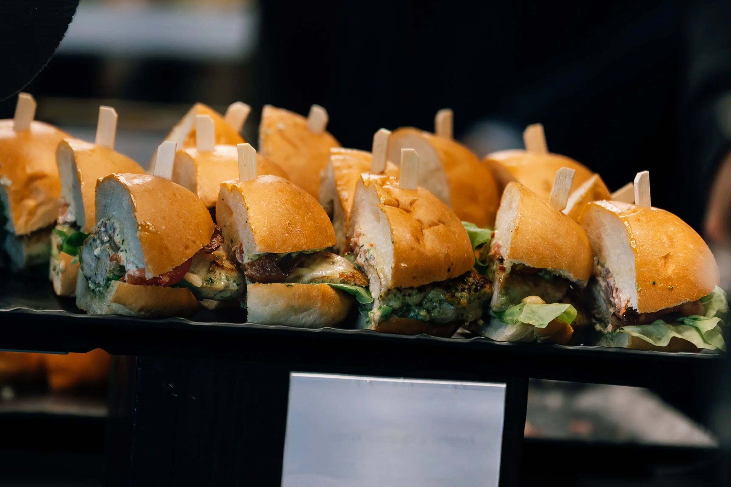 A close-up of a black tray of toothpick-skewered chicken sliders on the catering table.