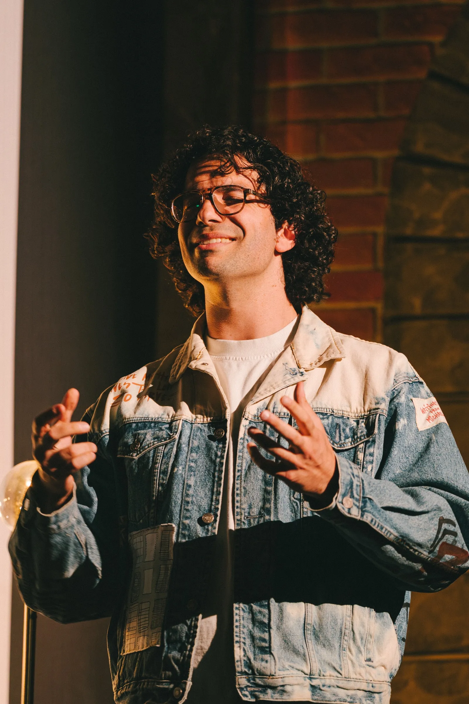 A storyteller in a patched denim jacket and glasses speaks with open hands, warmly lit against an exposed-brick wall.