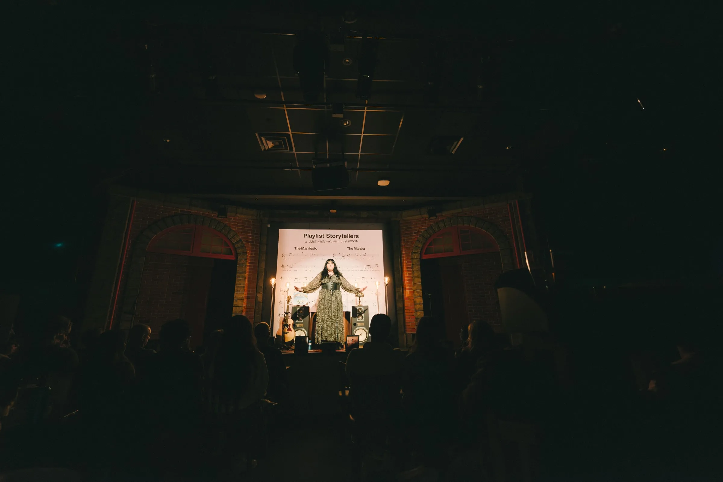 A wide, dark view of the theatre: a storyteller stands centre stage with arms outstretched under warm light, the “Playlist Storytellers” backdrop behind and the audience in silhouette.