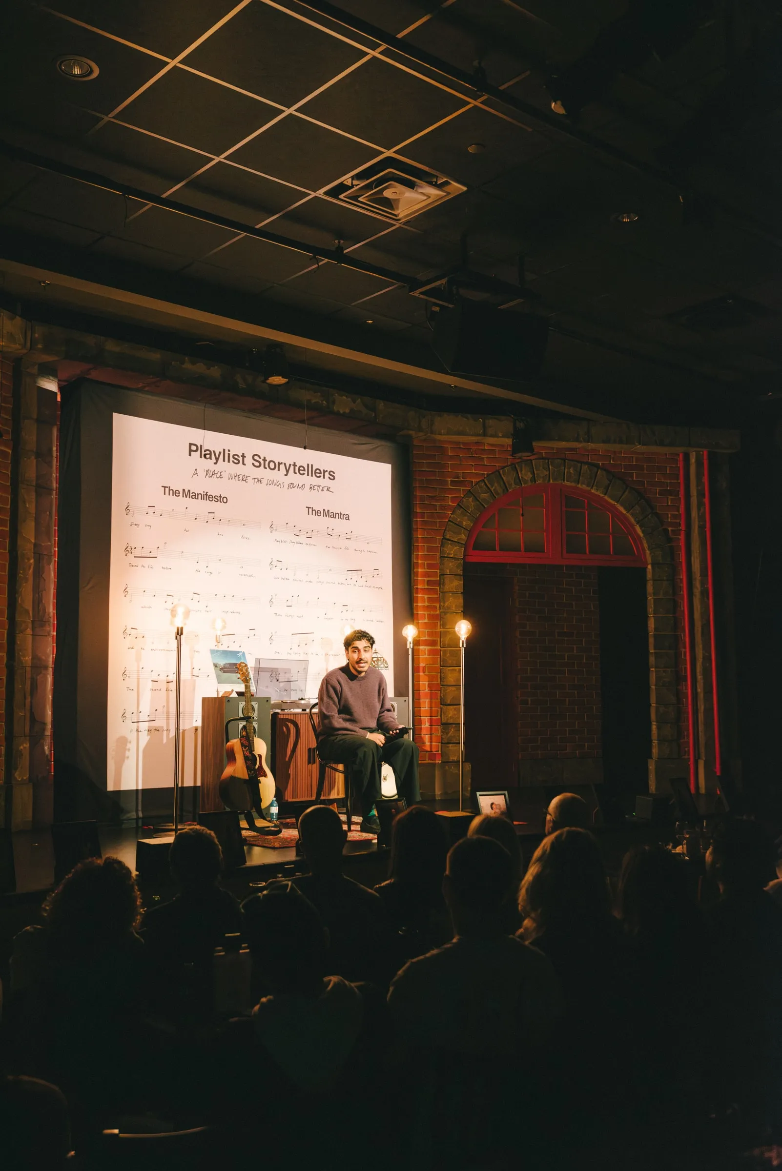A storyteller sits on a stool on the lit stage before a large projected “Playlist Storytellers” sheet-music backdrop, the audience silhouetted in the foreground.