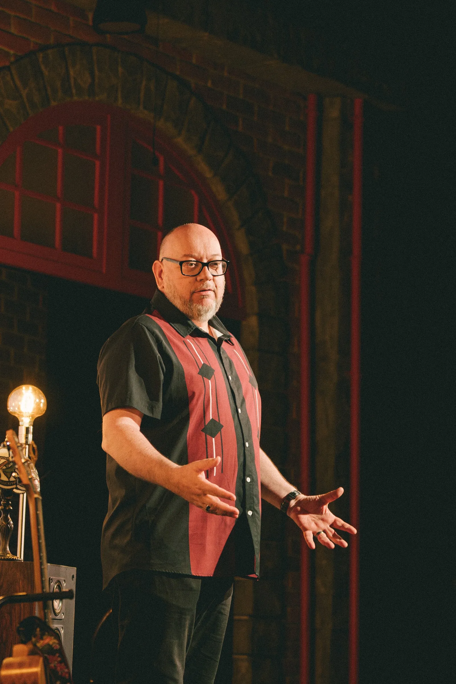 A bald storyteller in a red-and-black patterned shirt and glasses gestures mid-story beneath a red-framed brick archway.