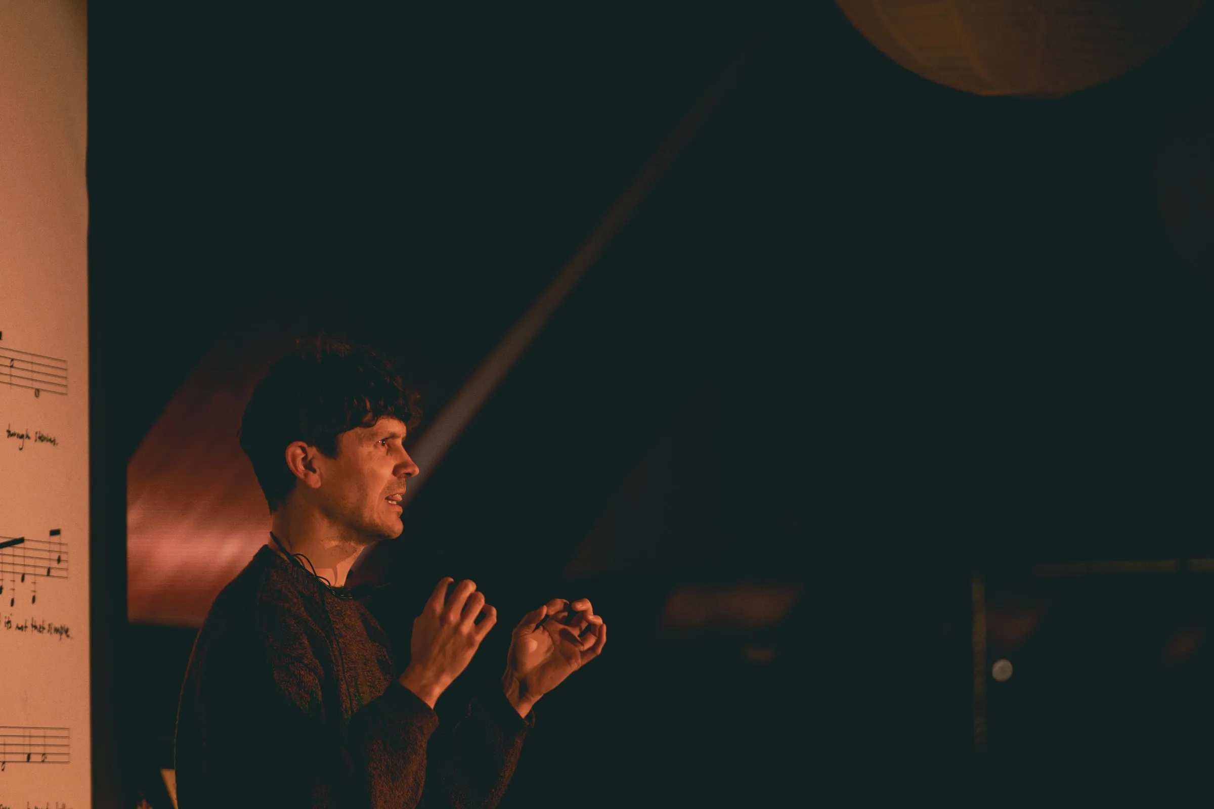 A storyteller in profile gestures with both hands as he speaks, warmly lit beside the printed sheet music.