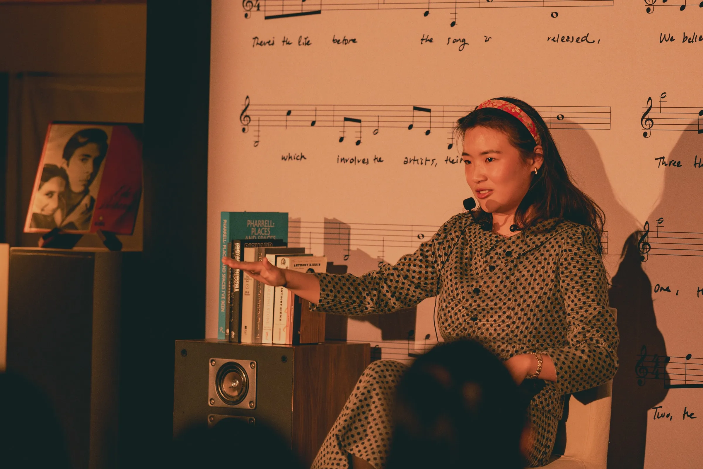A storyteller in a polka-dot outfit and red headband sits onstage gesturing toward a stack of books atop a speaker, in front of the printed sheet-music backdrop.