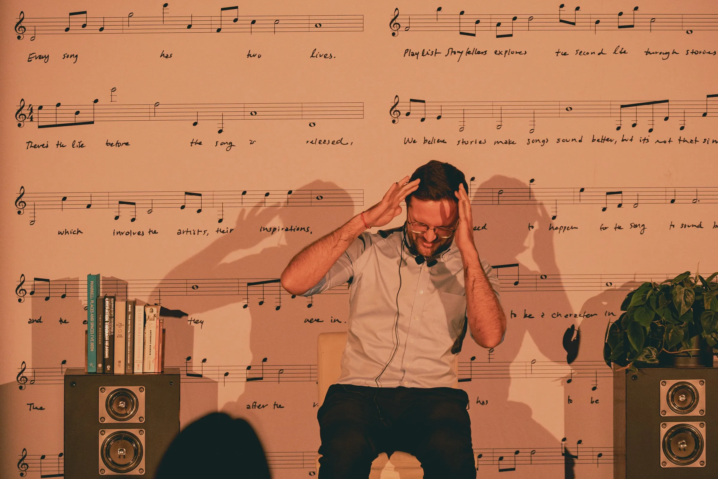 A storyteller sits on a stool with both hands pressed to his head mid-story, his shadow thrown large across the sheet-music wall behind him.