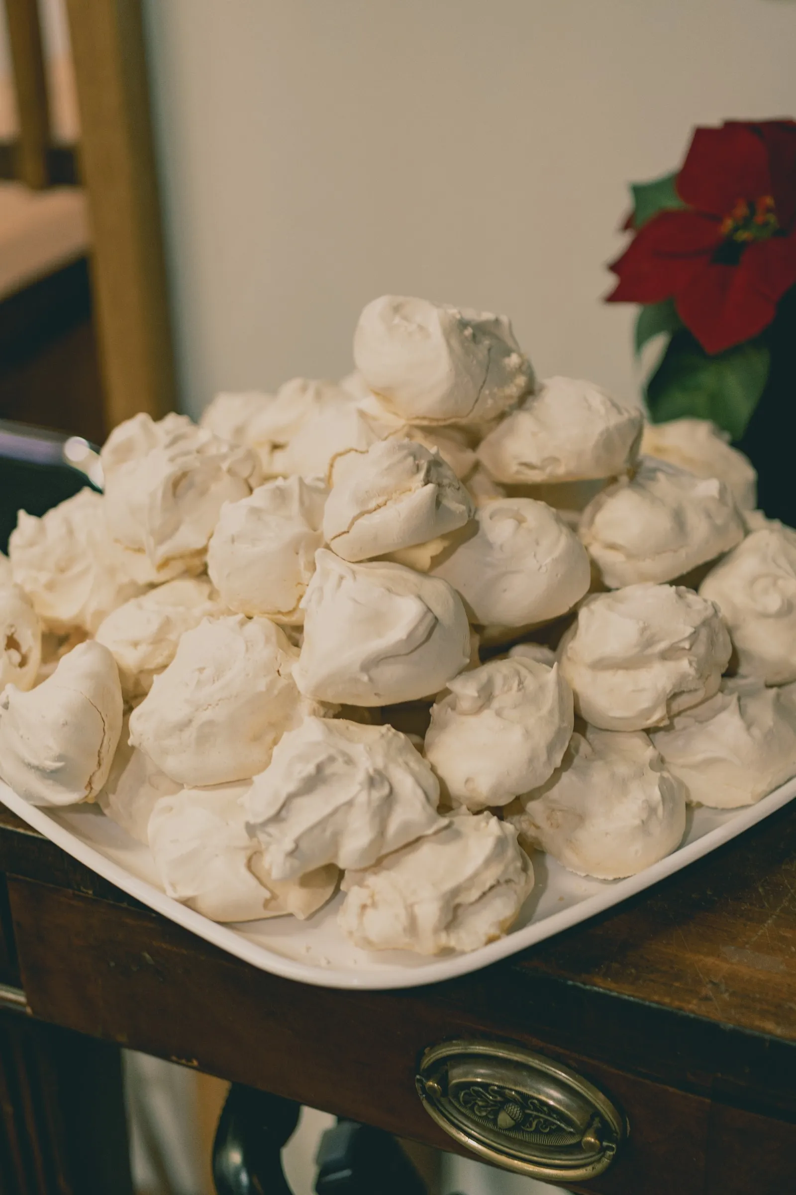 A white platter heaped with meringues on a wooden side table, a red poinsettia beside it.