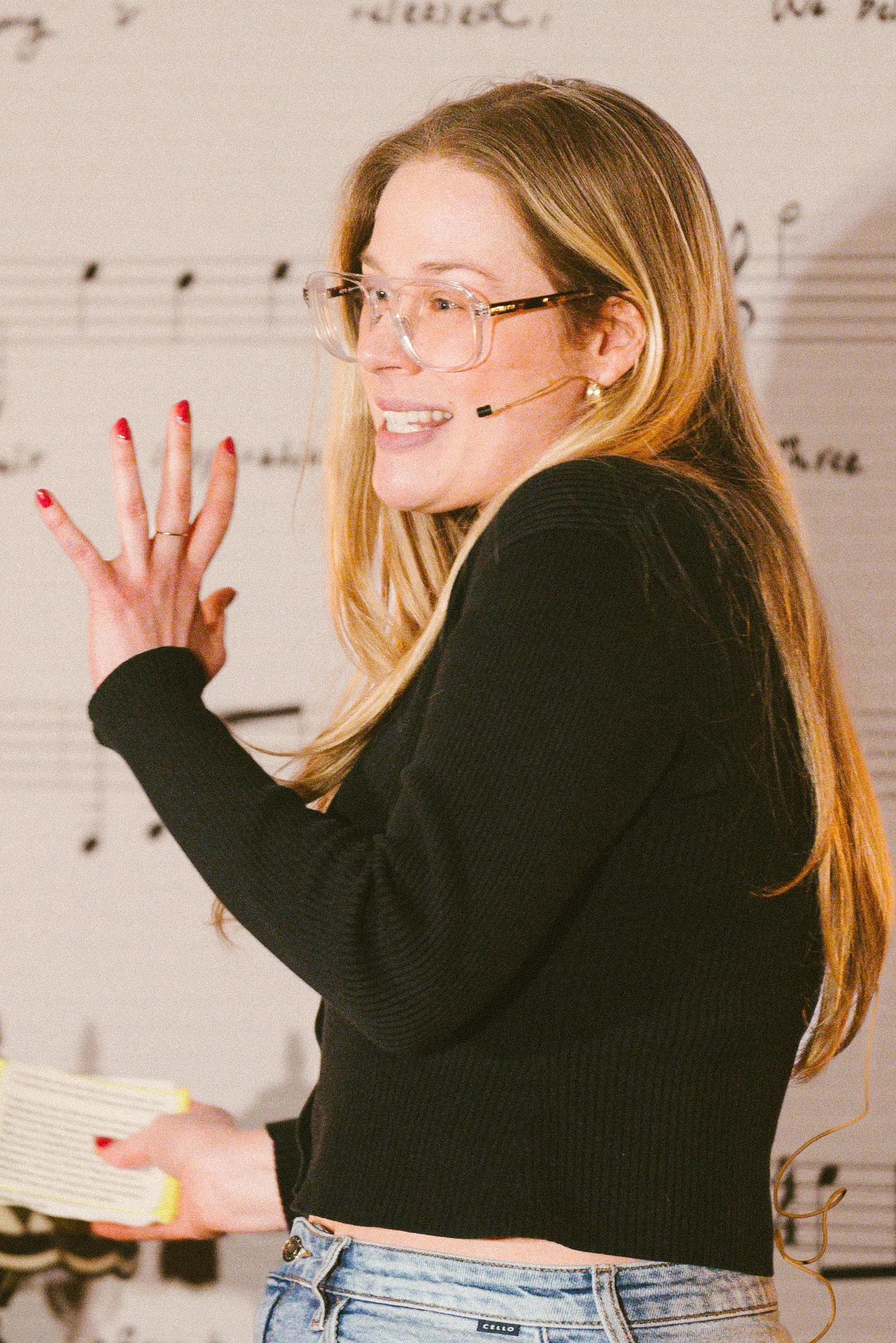 A storyteller in glasses and a headset microphone glances back over her shoulder mid-gesture against the wall of printed sheet music.