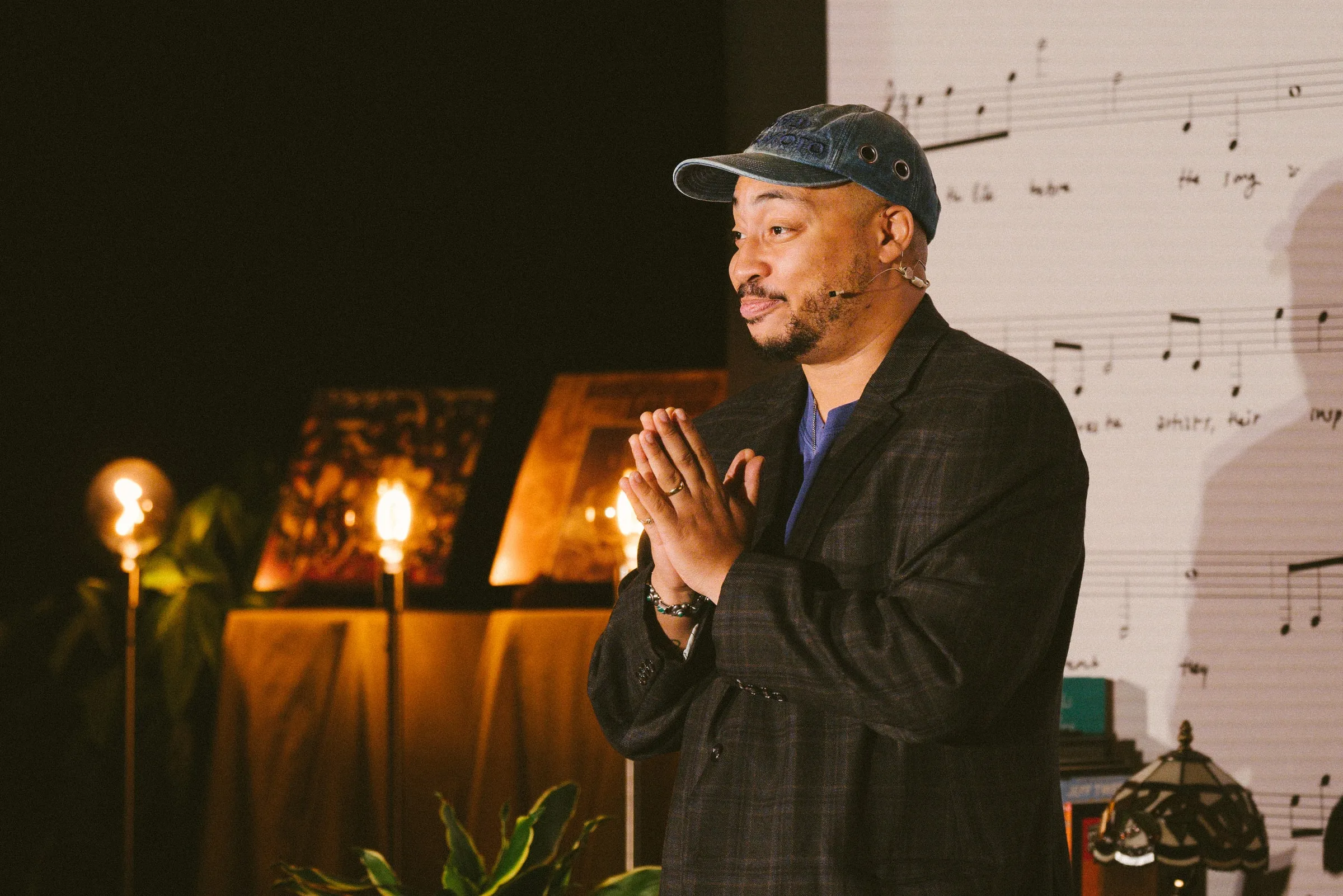 A storyteller in a checked blazer and ball cap stands with hands pressed together, lit by warm lamps in front of the printed sheet-music backdrop.