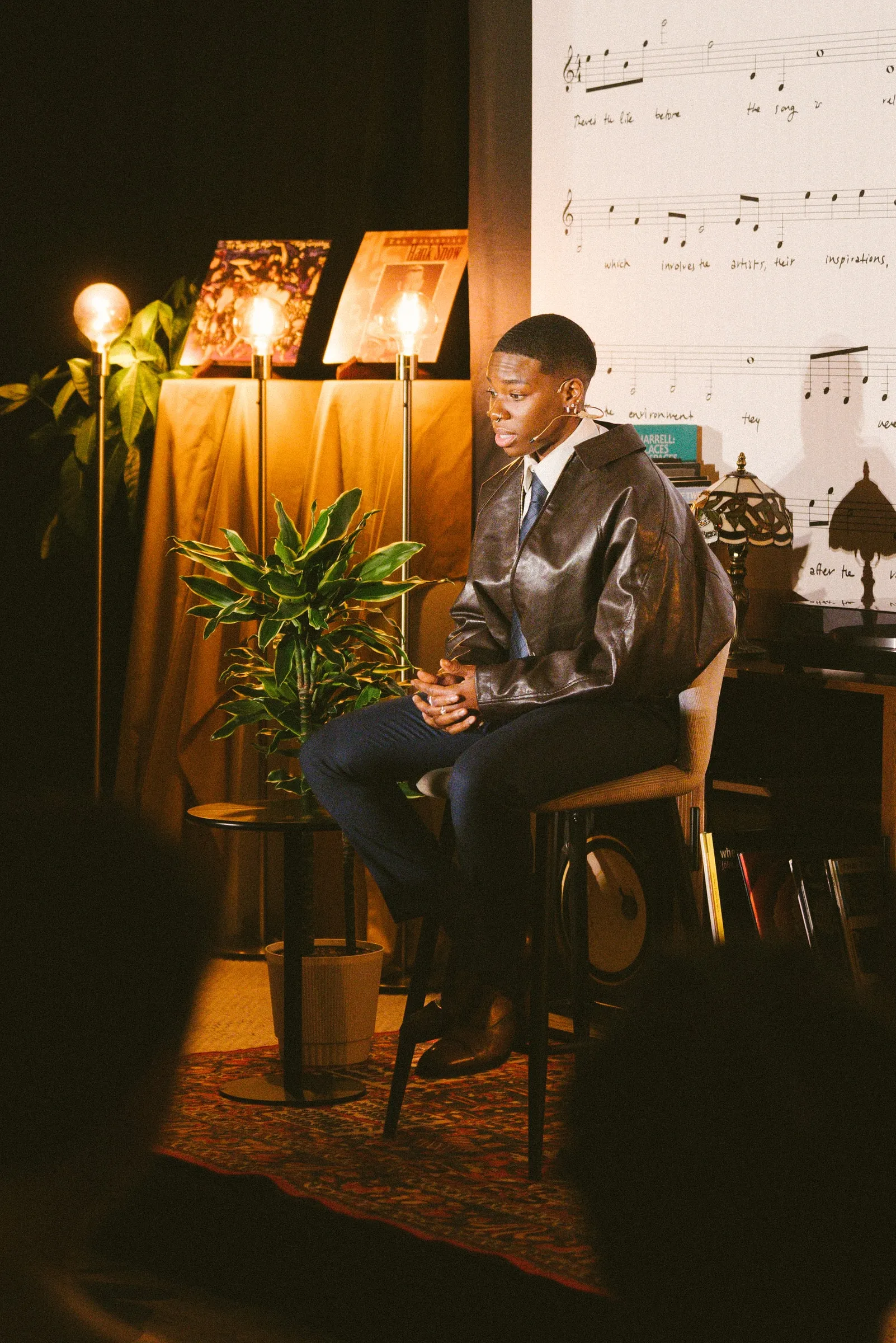 A storyteller in a leather jacket sits on a stool under warm lamplight, a potted plant beside him, before the sheet-music backdrop.