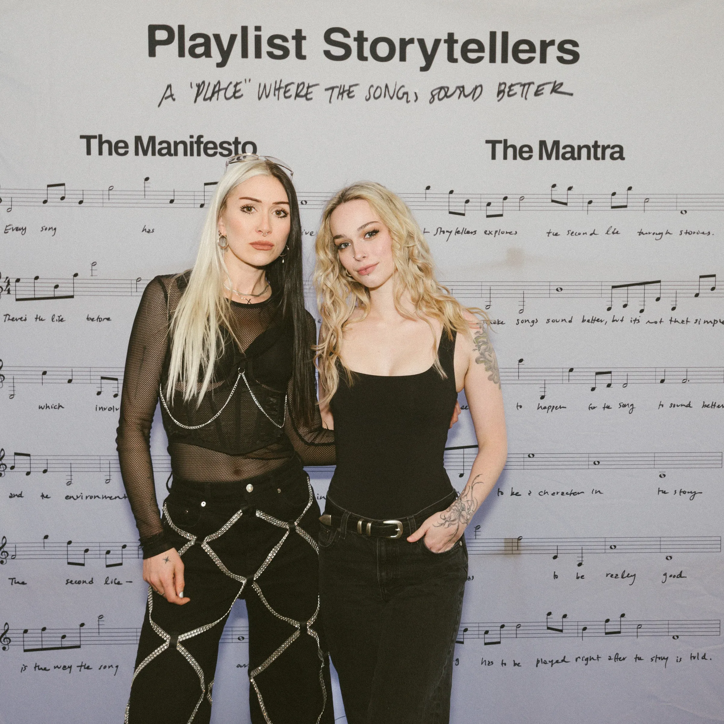 Two guests pose together at the step-and-repeat backdrop printed “Playlist Storytellers” over sheet music.