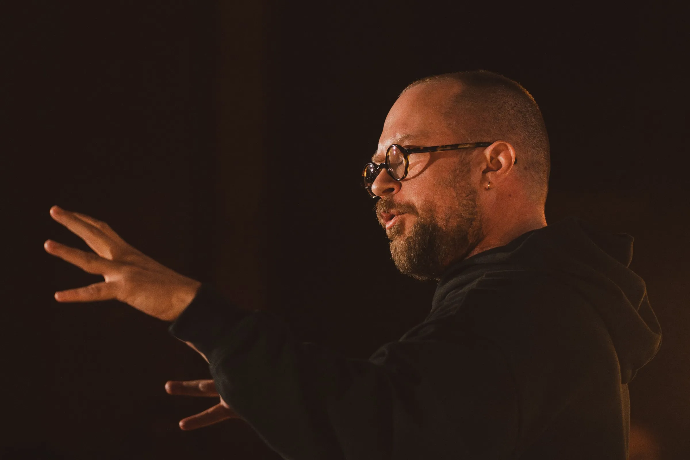 A bearded storyteller in glasses and a dark hoodie gestures with both hands mid-story, warmly lit against the dark room.