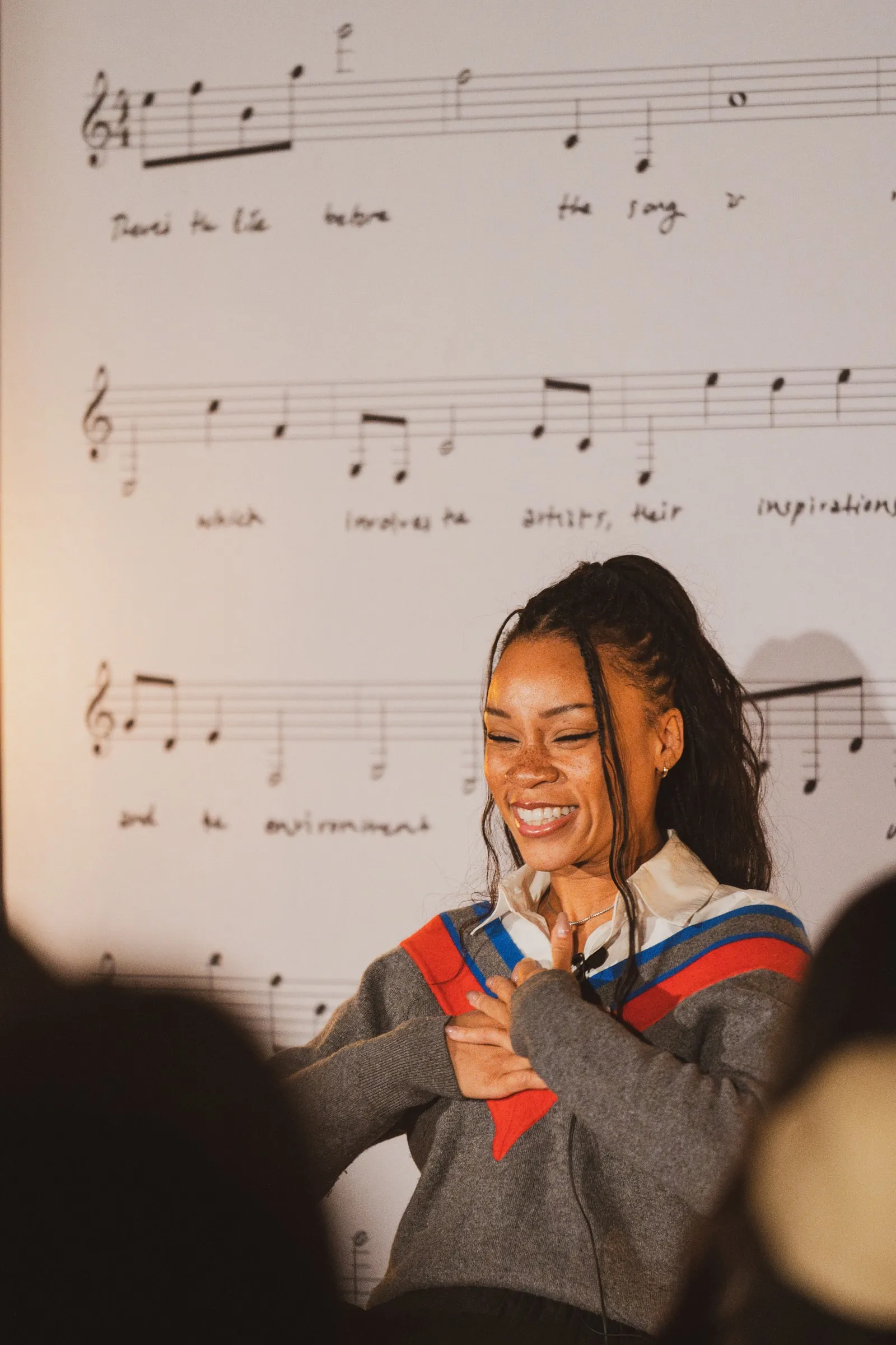 A storyteller in a grey-and-red varsity cardigan smiles with hands clasped to her chest in front of the oversized printed sheet music.