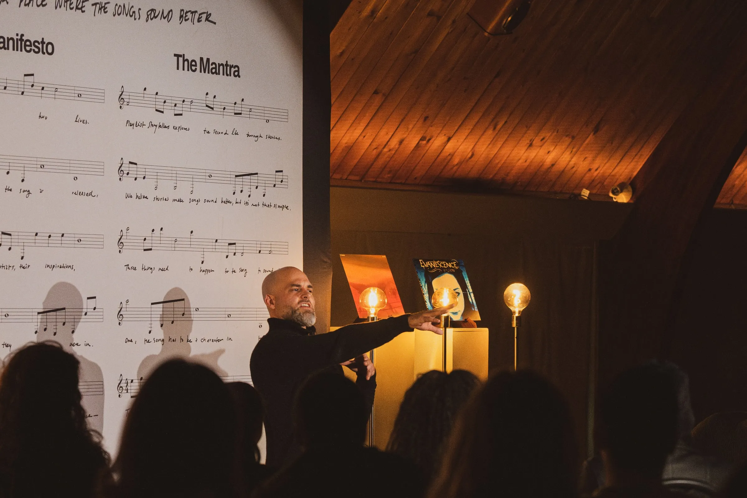 A storyteller reaches toward a glowing record display on plinths beneath the printed sheet-music backdrop, the audience silhouetted under the venue’s warm wooden ceiling.