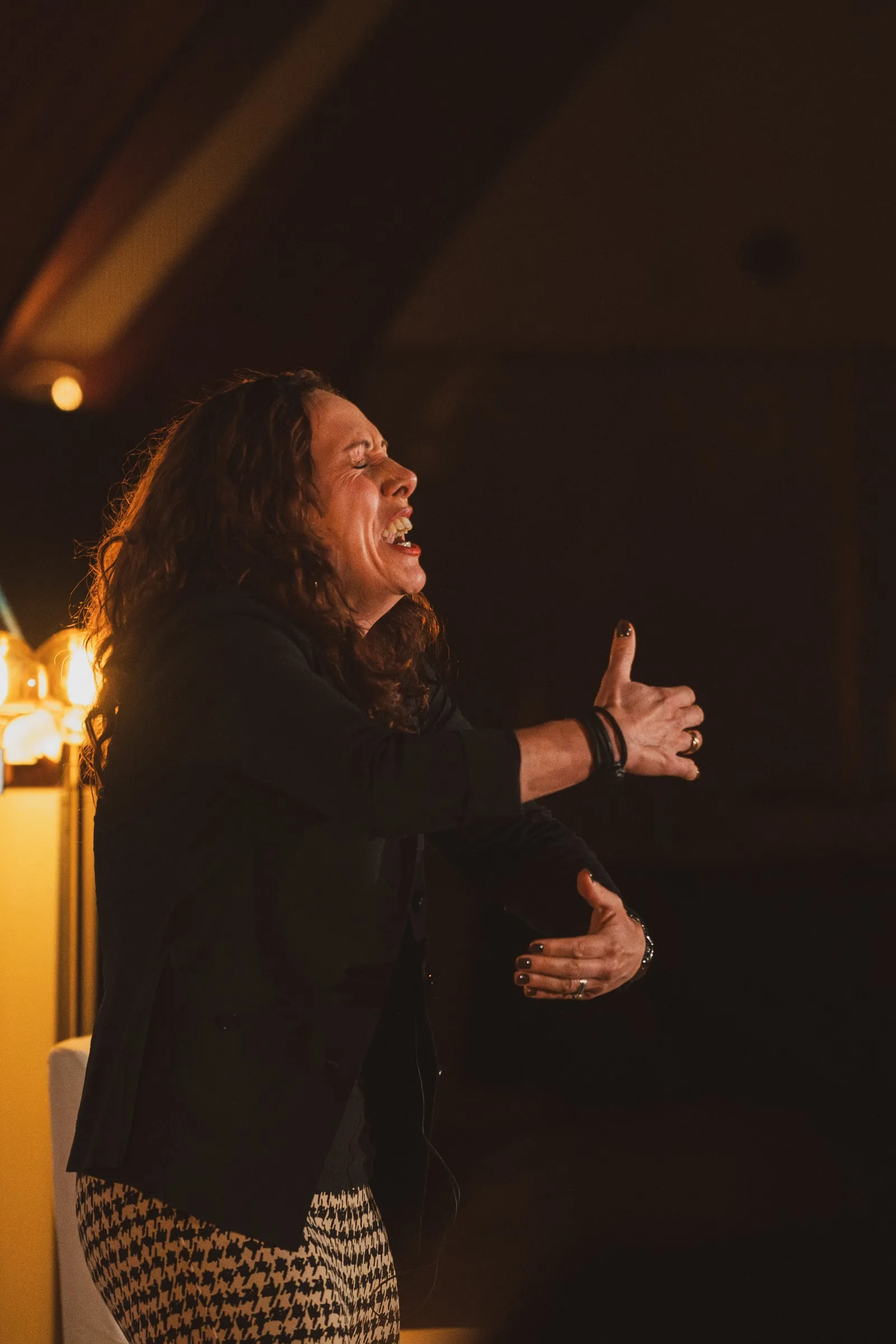 A storyteller in a black blazer throws her head back laughing, hands open, lit warm against the darkness.