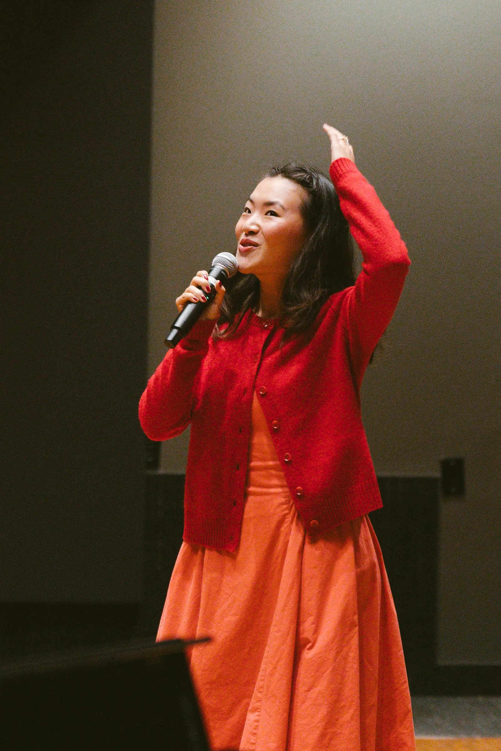 The speaker, in a red cardigan, addresses the room with a handheld microphone, one arm raised mid-gesture.