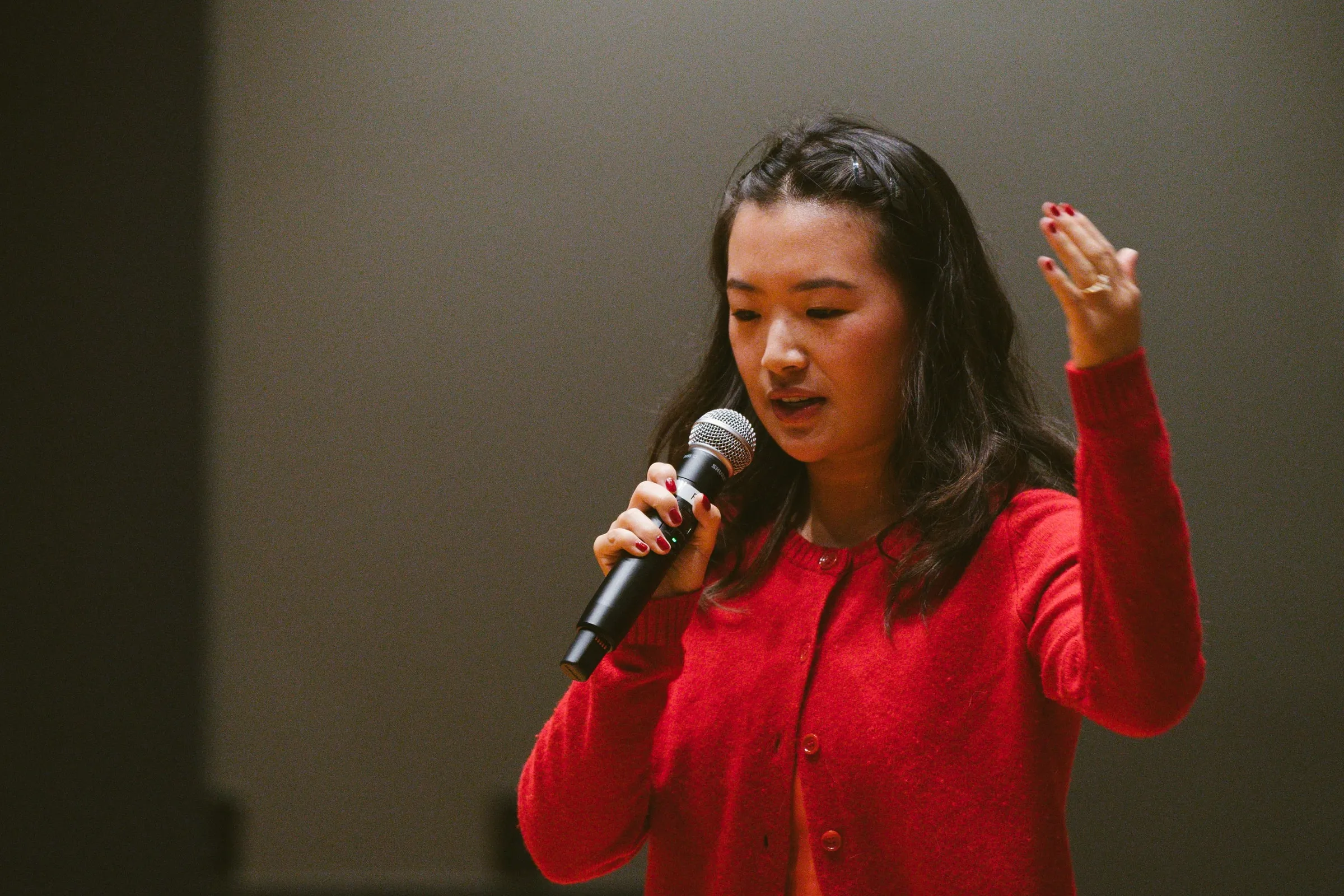 The speaker, in a red cardigan, speaks into a handheld microphone with eyes lowered in thought, a hand raised.