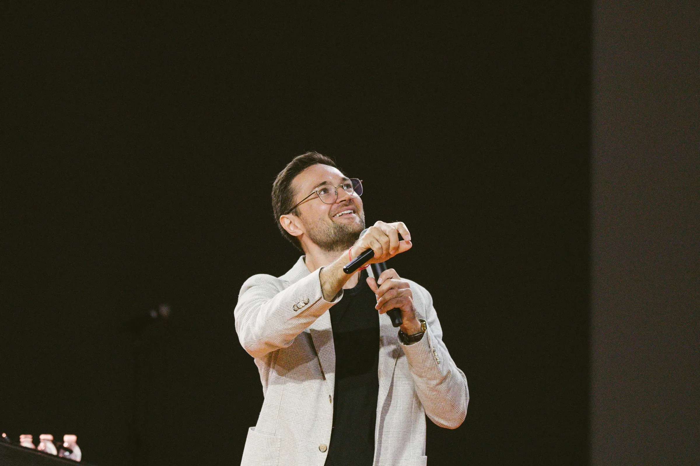 The speaker, in a light checked blazer, holds a handheld microphone and looks upward with a smile.
