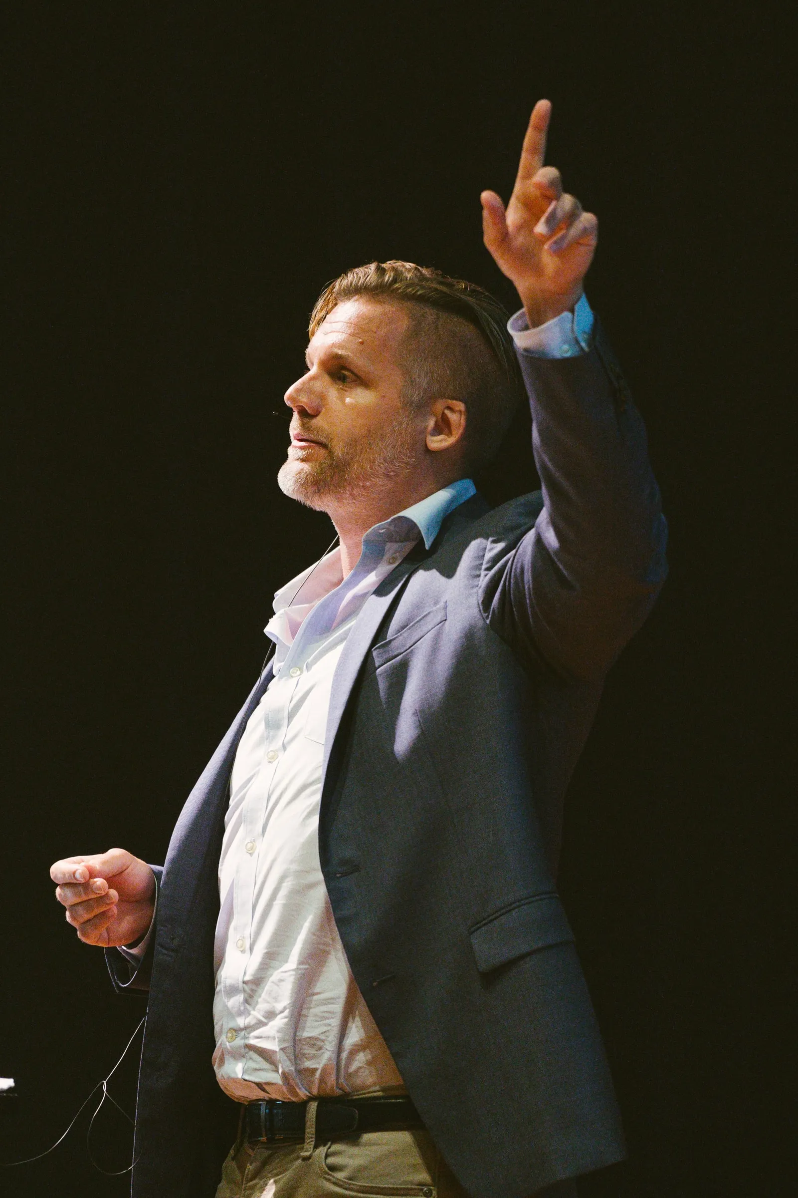 The speaker, in a grey blazer, raises a finger to make a point, lit against a black backdrop.