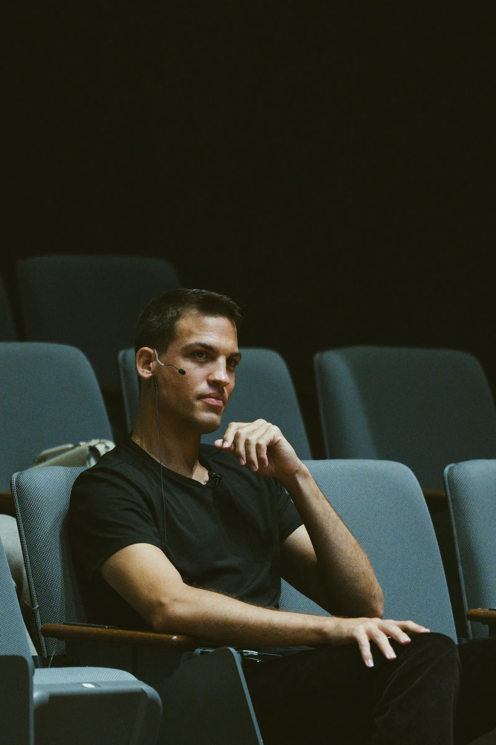 The speaker sits alone in the theatre seats before the talk, a headset microphone on, resting his chin on his hand.