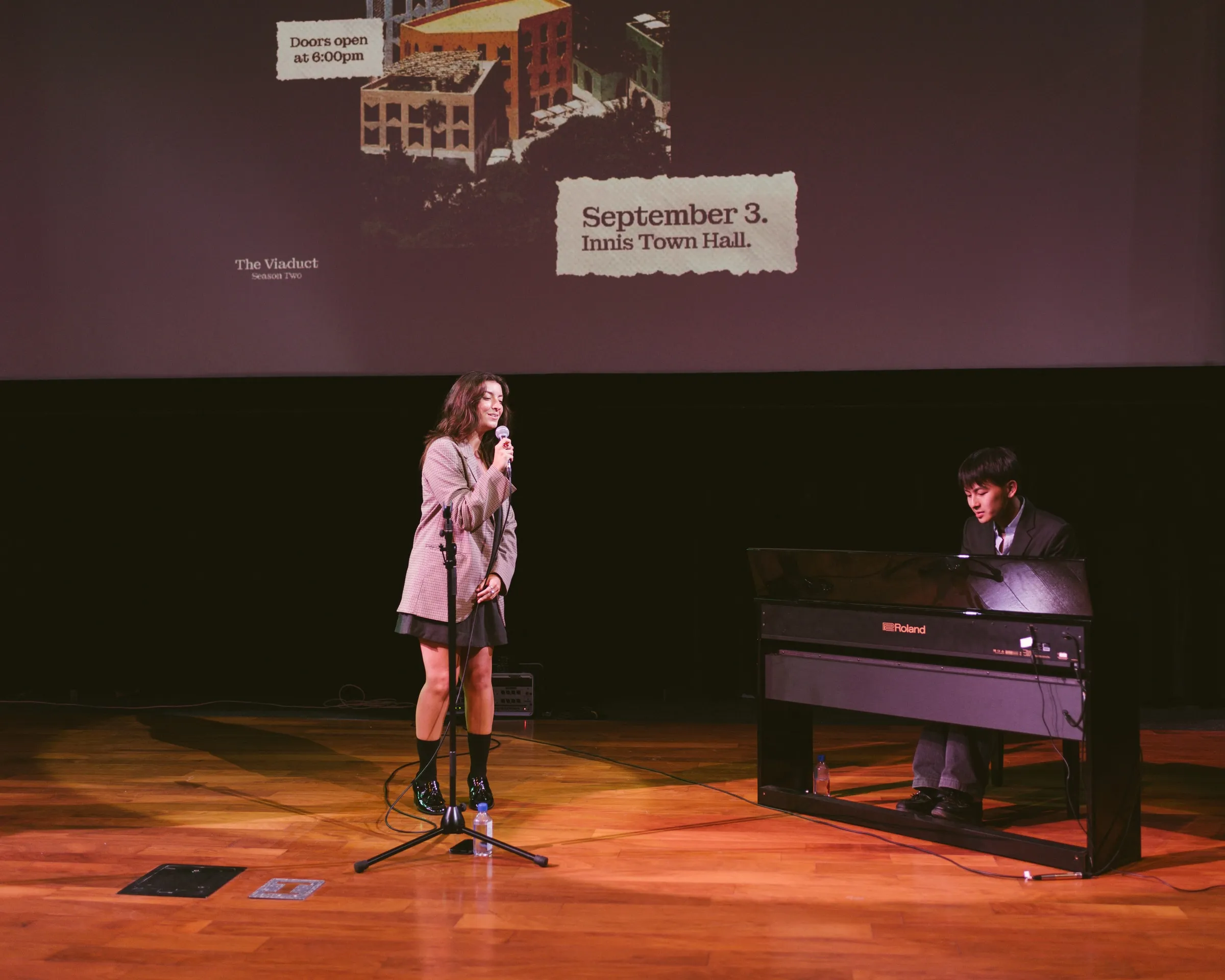 A singer performs into a standing microphone while a pianist accompanies on a keyboard, beneath the projected Viaduct title slide for the September 3 talk at Innis Town Hall.