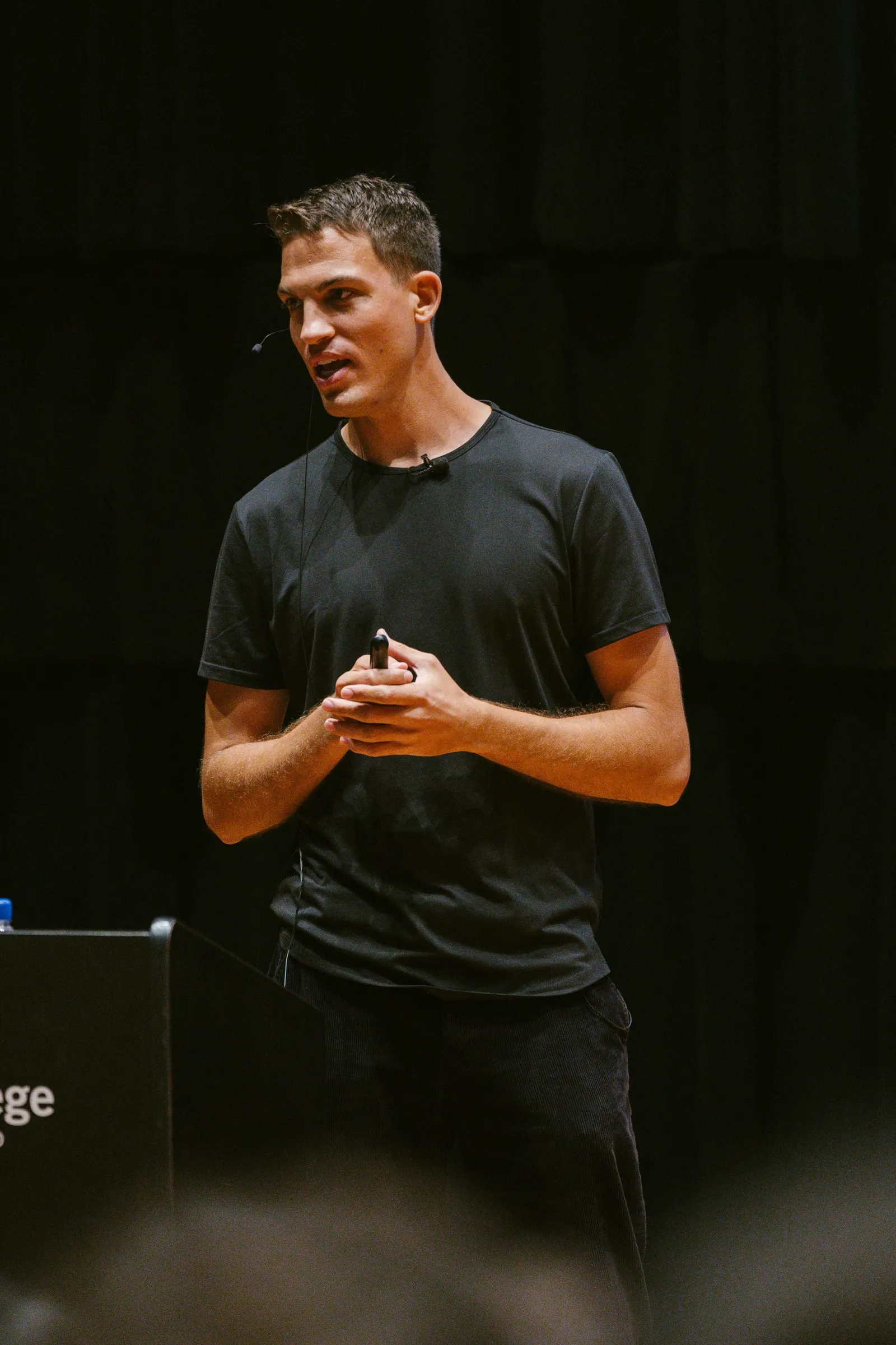 The speaker, in a black T-shirt and headset microphone, stands mid-lecture holding a presentation clicker against a dark stage backdrop at Innis Town Hall.