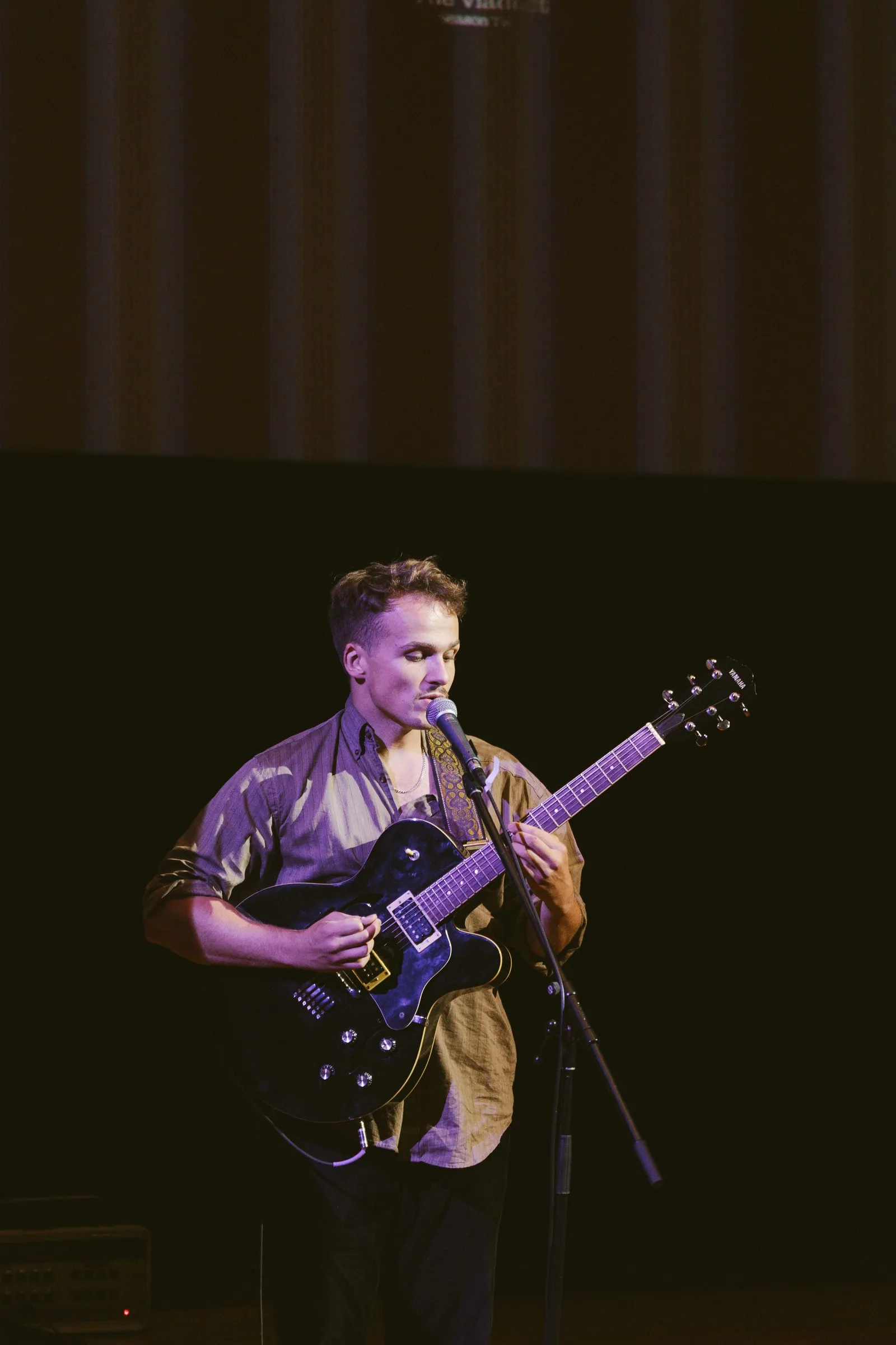 A musician plays a hollow-body electric guitar and sings into a microphone on the dimly lit stage.