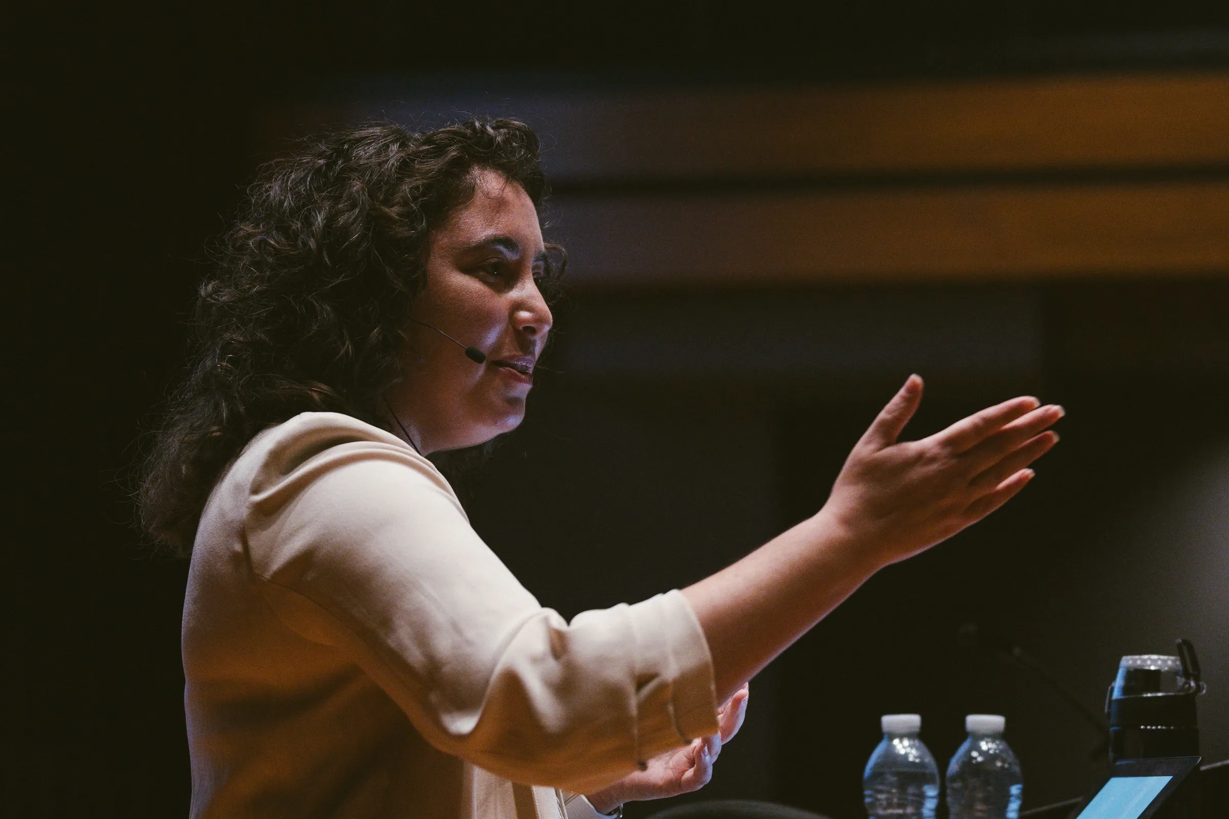 The speaker, in a cream blazer and headset microphone, gestures with an open hand mid-talk at the Innis Town Hall lectern.