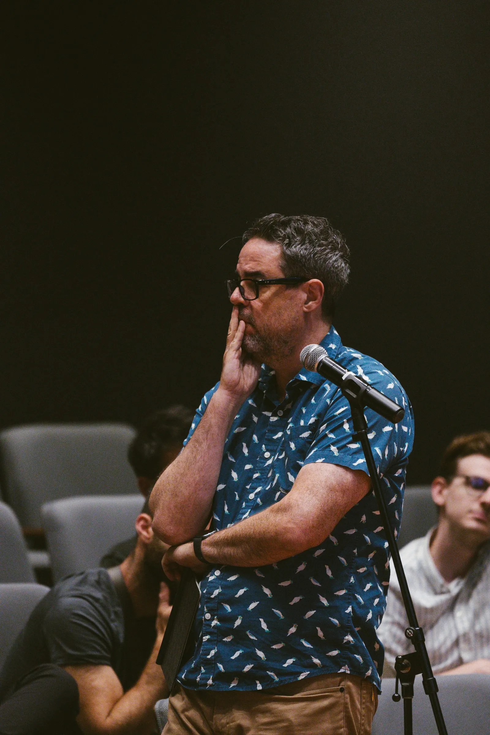 An audience member stands at a floor microphone, hand to his chin, to ask a question.