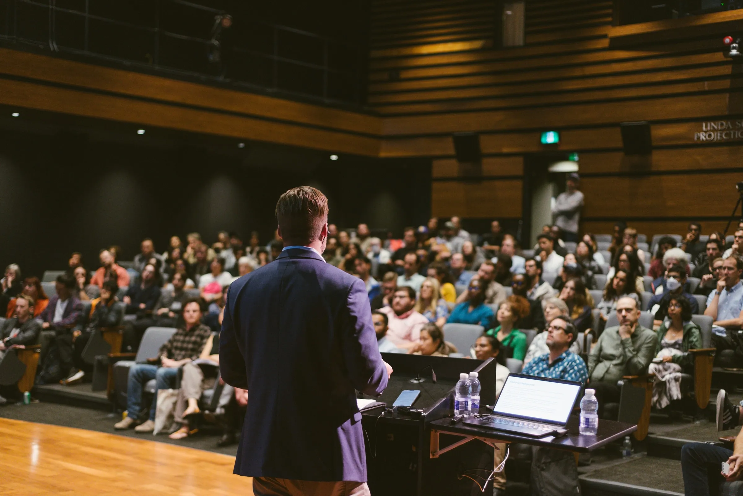 Seen from behind the podium, the speaker in a navy blazer faces a full, tiered audience in the wood-panelled Innis Town Hall auditorium.