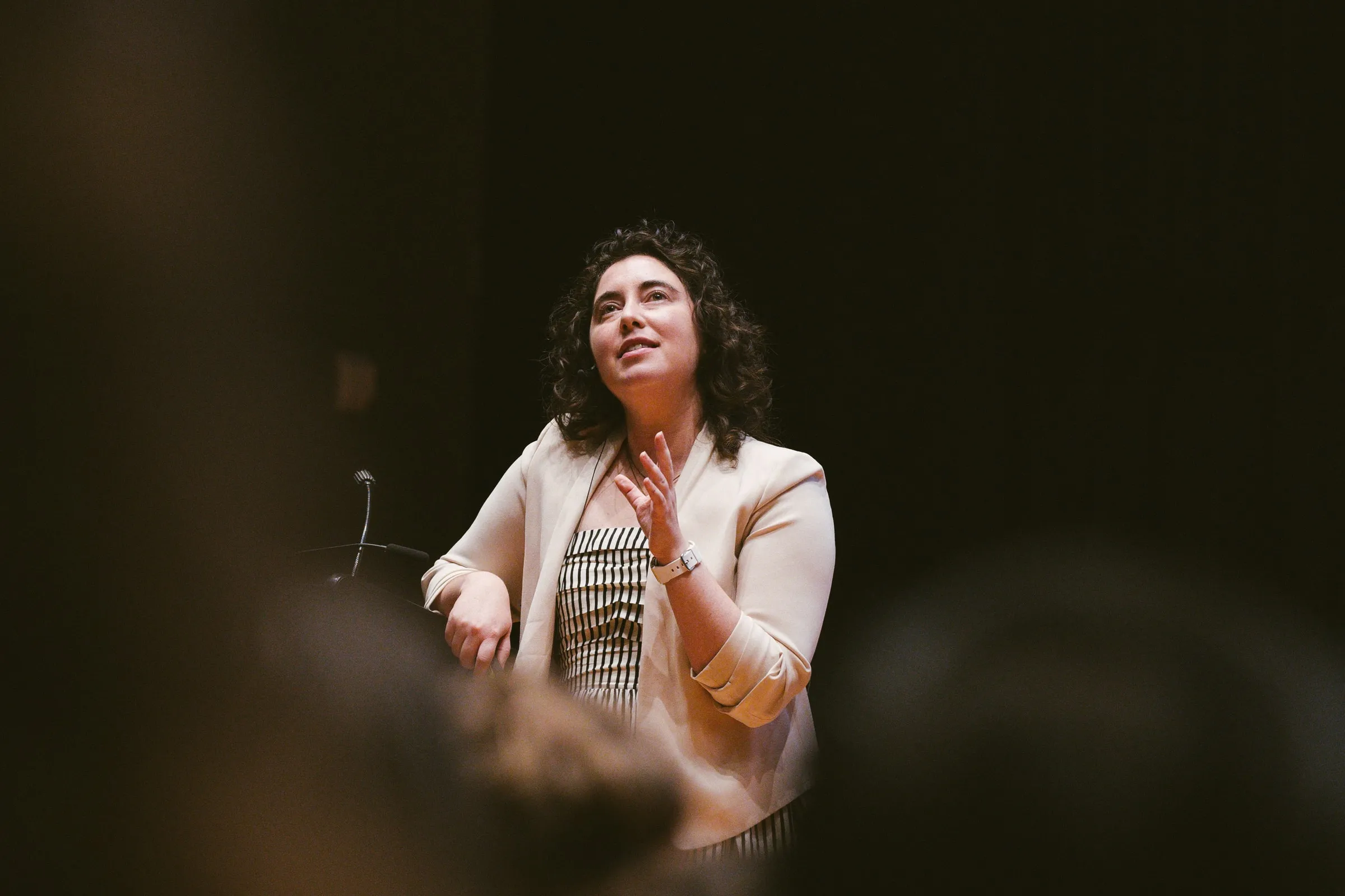 The speaker, in a cream blazer and headset microphone, stands at the lectern with a hand to her chest, glimpsed between the silhouetted heads of the darkened audience.