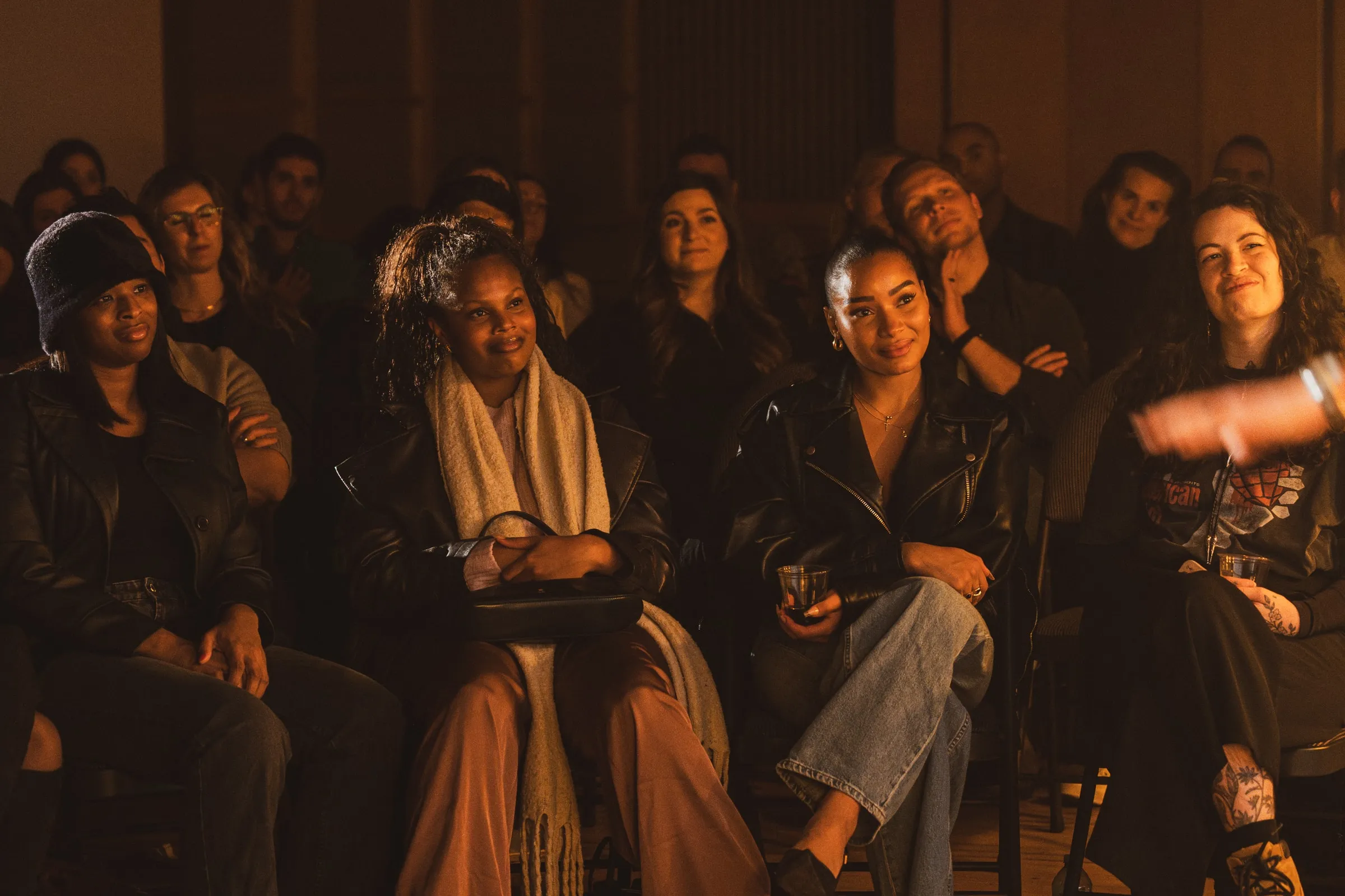 Audience members sit watching in warm lamplight, a front row of guests in leather jackets and a wool scarf smiling toward the stage at the 918 Bathurst show.