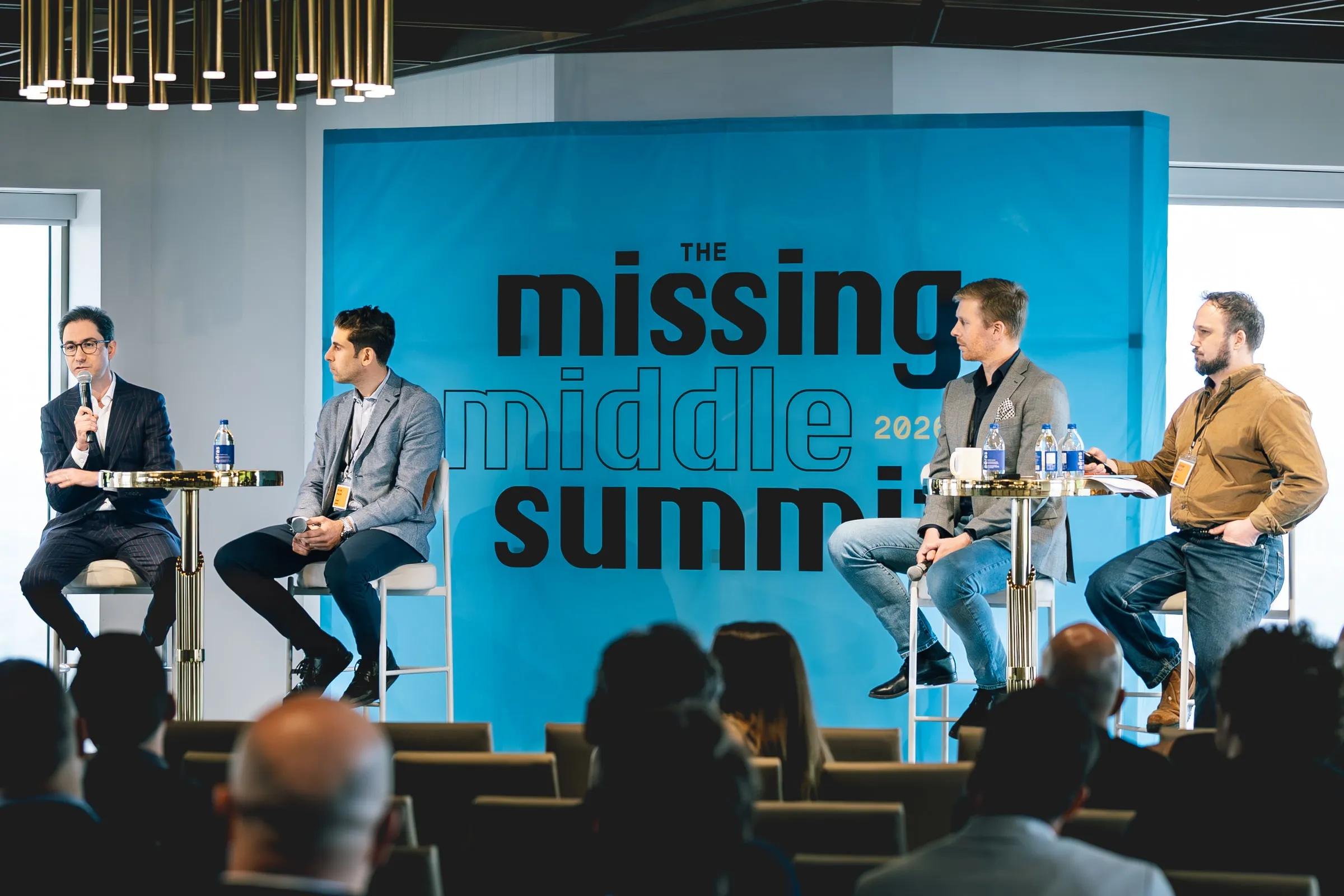 Four panelists sit on stools onstage, one speaking into a microphone, before a large blue backdrop reading “the missing middle summit 2026,” the audience silhouetted in the foreground.