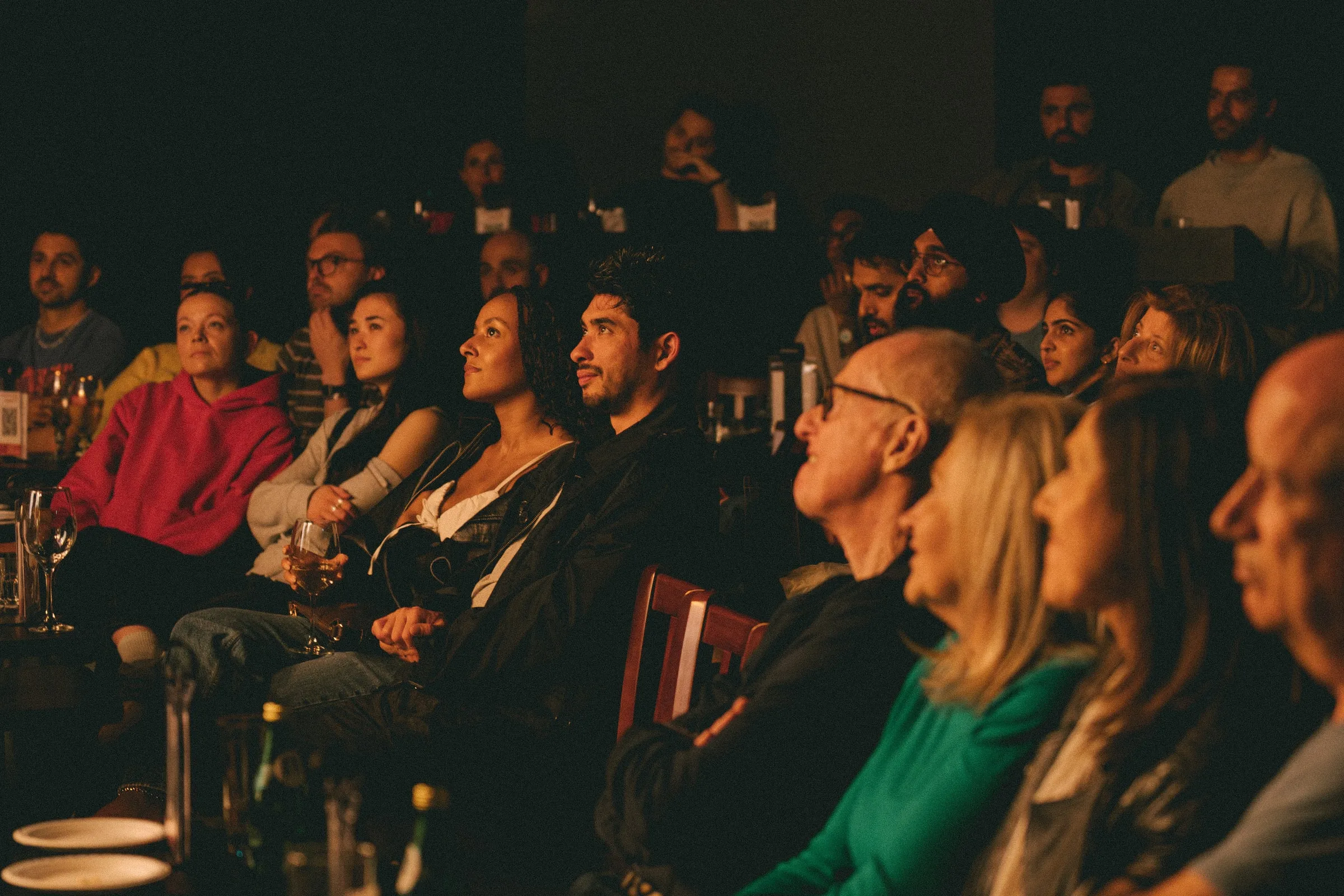 A warmly lit audience watches from rows of seats with drinks on the tables, faces turned toward the stage at the Second City show.