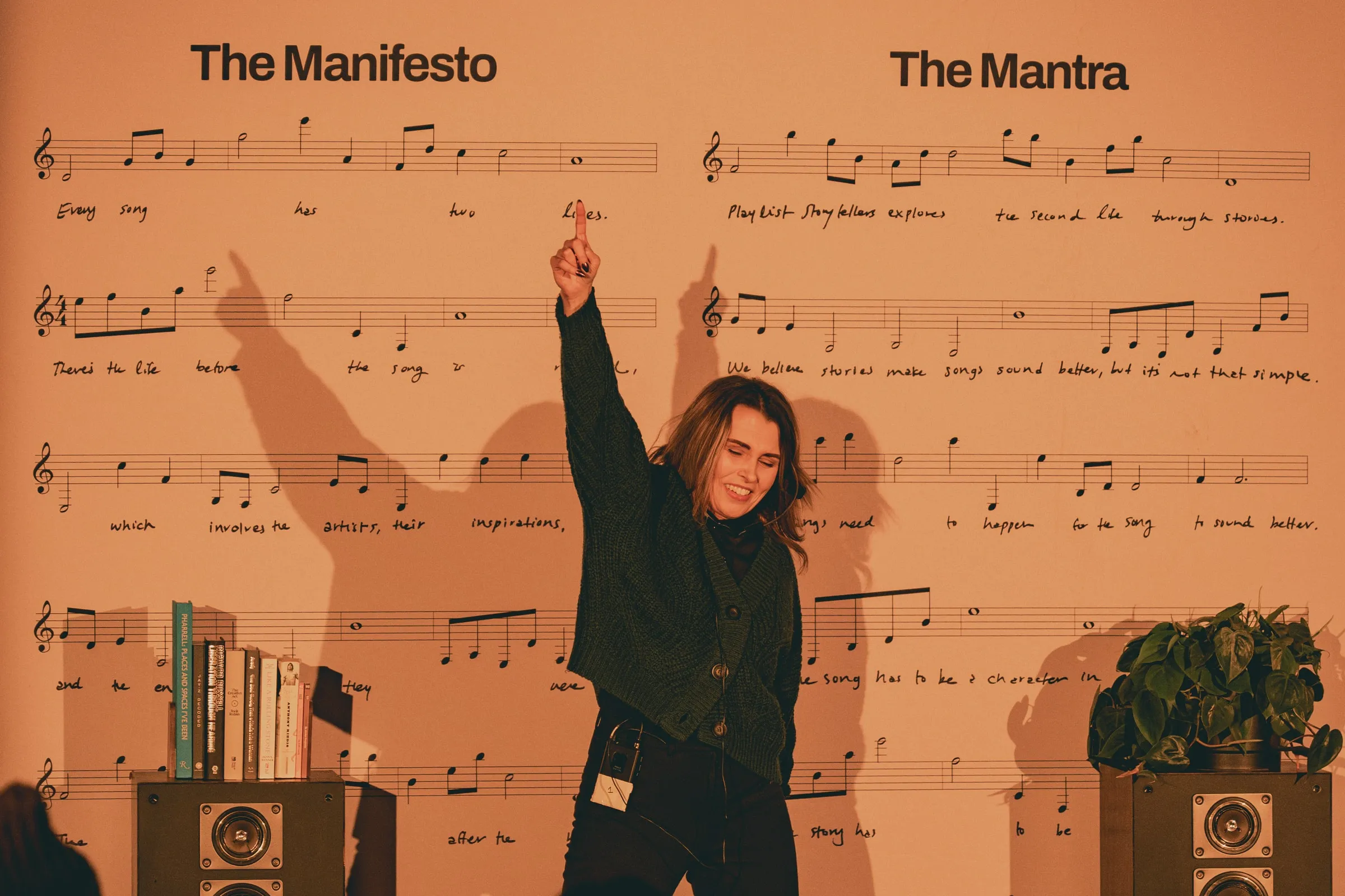 A storyteller in a dark green cardigan throws one arm overhead, her shadow cast on the wall of printed sheet music headed “The Manifesto” and “The Mantra,” with books and a plant resting on speakers beside her.