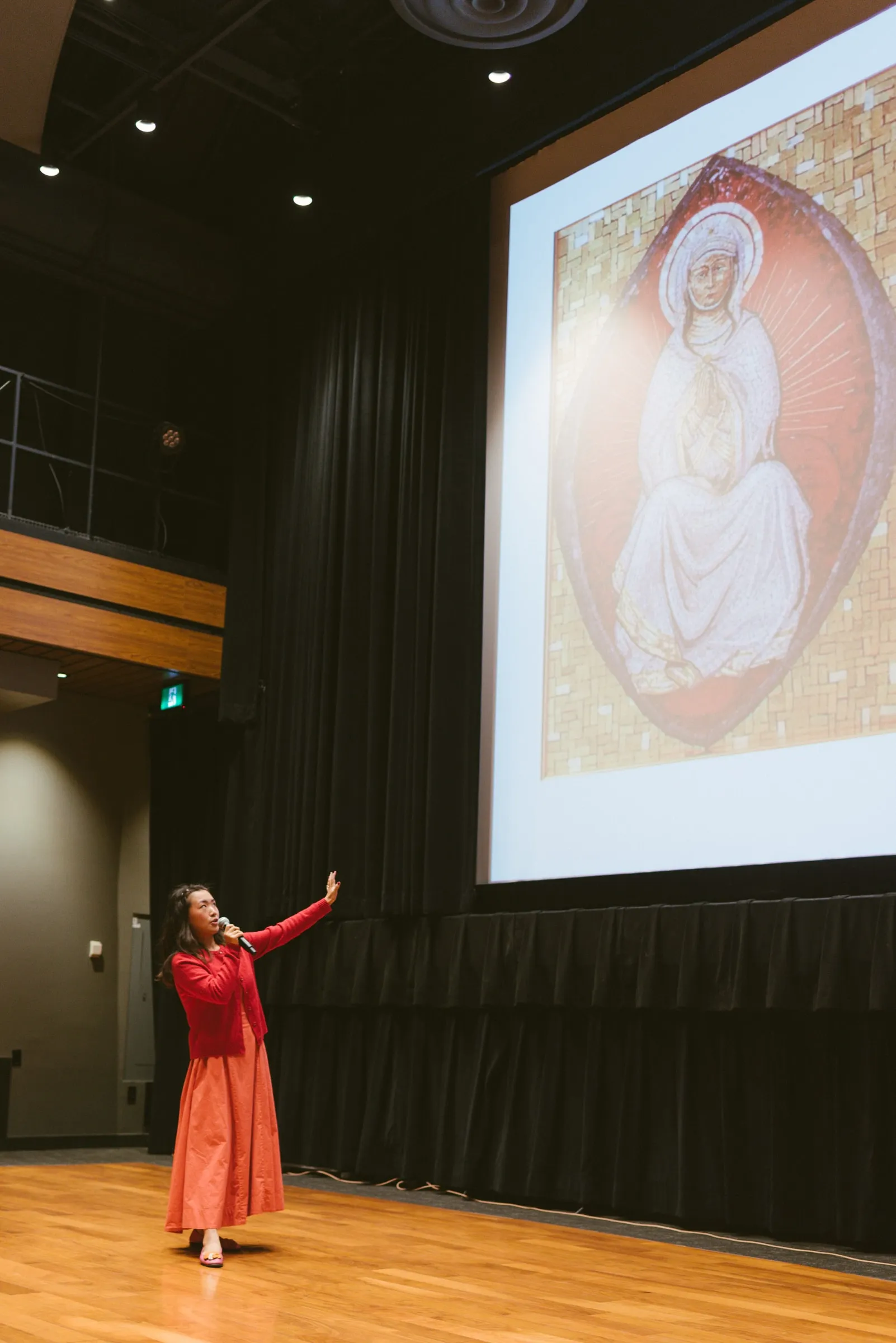 The speaker, in a red cardigan and long coral skirt, stands on the wooden stage with one arm raised toward a large projected mosaic of a haloed seated figure, against the black drapes of Innis Town Hall.