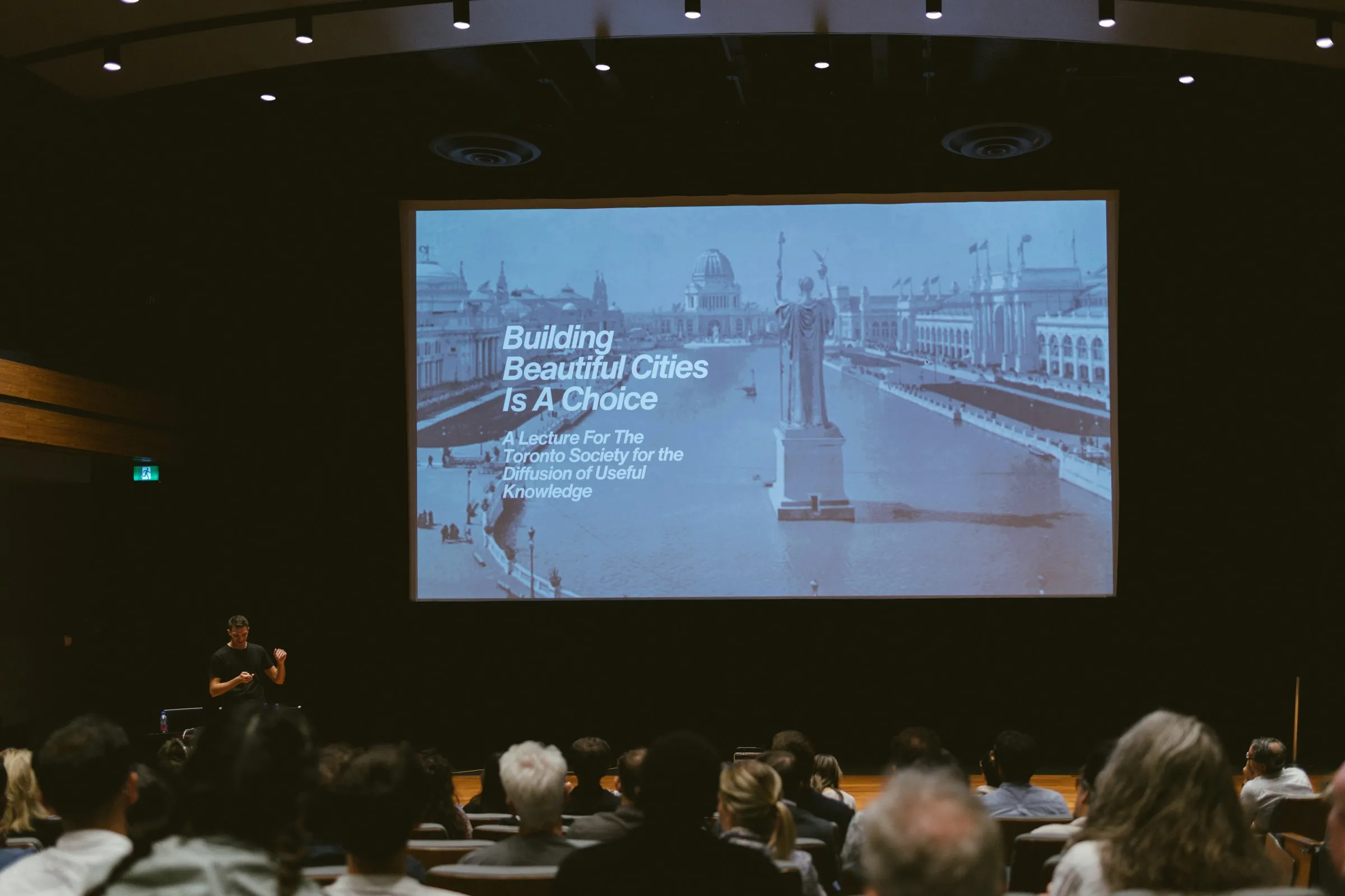 A speaker stands small at the foot of the large Innis Town Hall screen, which shows a classical waterfront cityscape and the title slide “Building Beautiful Cities Is A Choice,” the silhouetted audience filling the foreground.