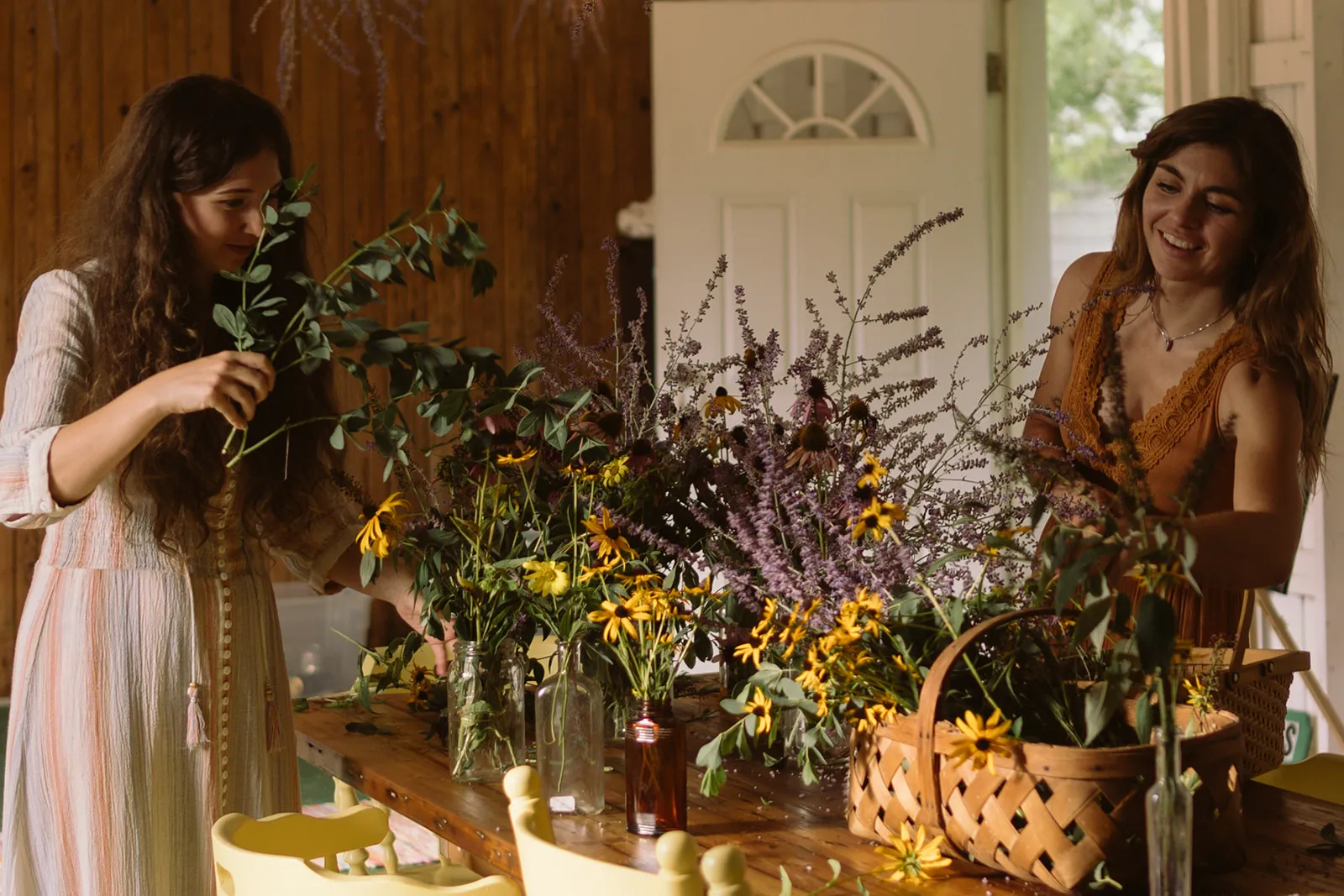 Two women arrange wildflowers — black-eyed Susans, lavender, and eucalyptus — in glass jars along a long wooden table, one holding a sprig of eucalyptus to her face, in a green-panelled room with an open white door behind them.