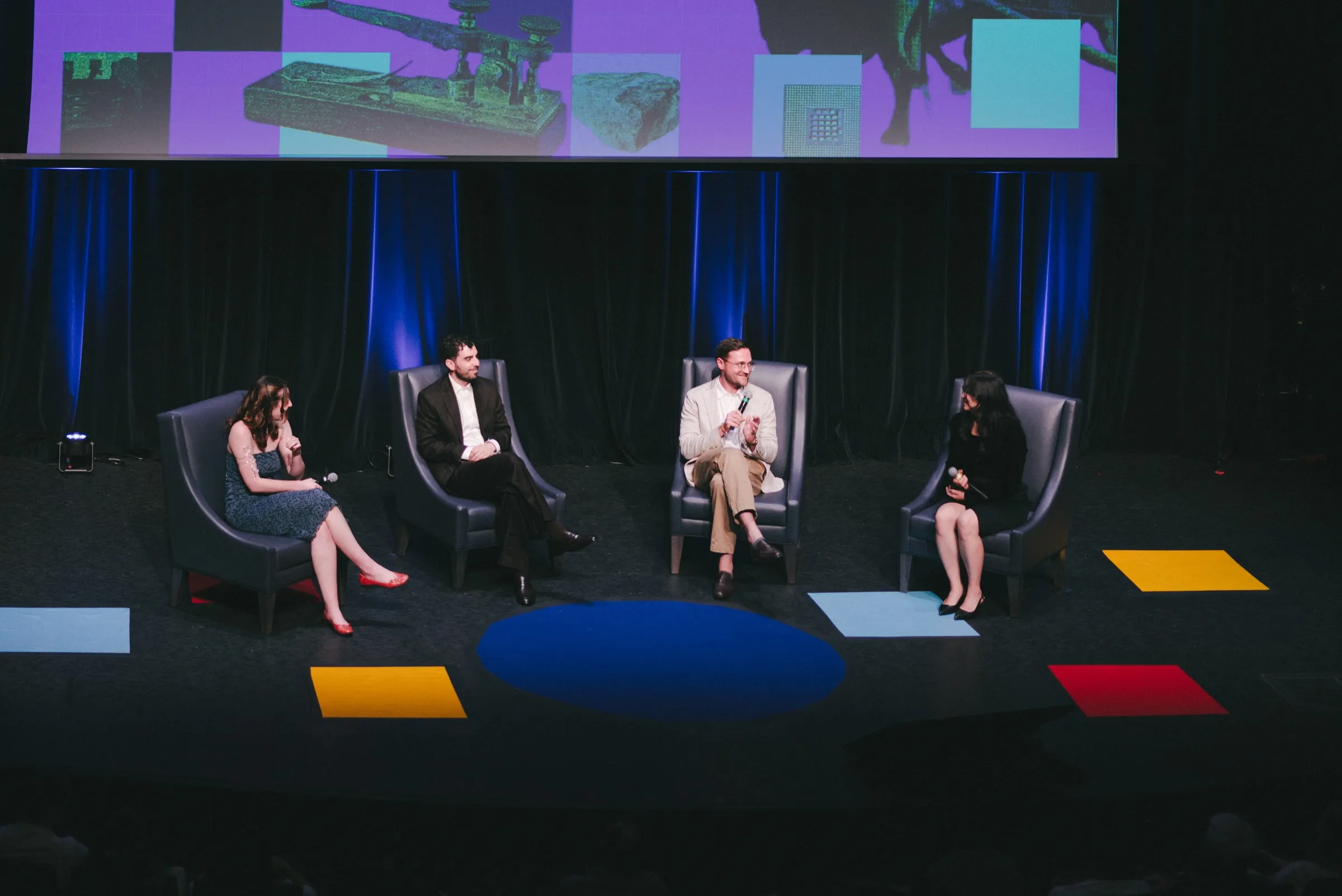 Four people sit in armchairs onstage in conversation, microphones in hand, on a set scattered with colourful squares and a blue oval, beneath a large screen of green-toned images at the Isabel Bader Theatre.