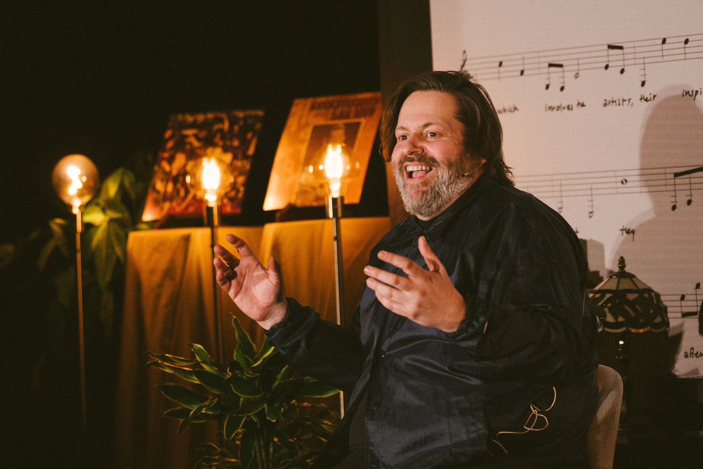 A bearded storyteller in a navy shirt sits gesturing with both hands and laughing, beside a draped table of framed album covers lit by warm Edison-bulb lamps in front of the printed sheet-music backdrop.